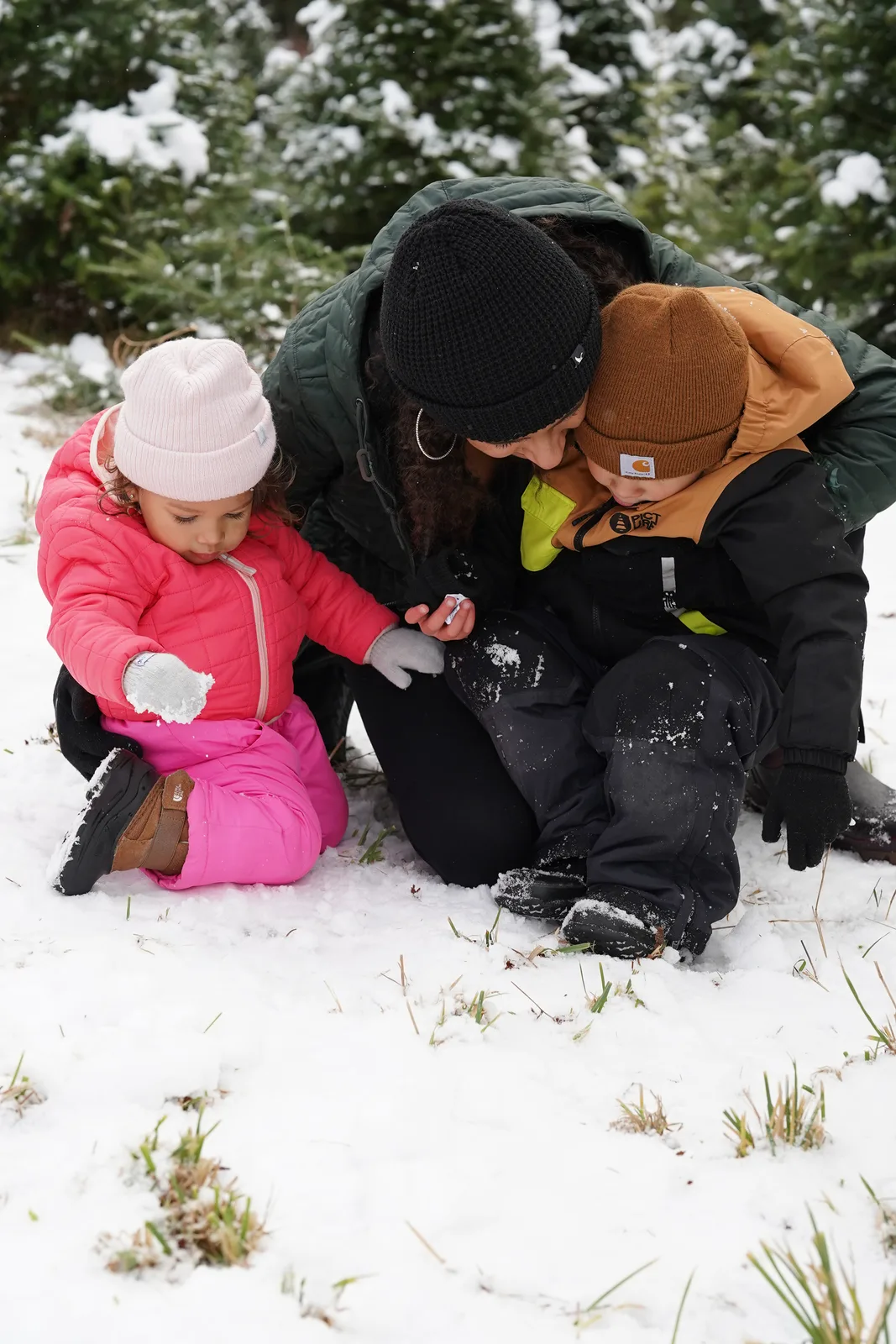 Adult in black beanie and green jacket with child in pink snowsuit and light hat and child in brown beanie and black jacket on snow.