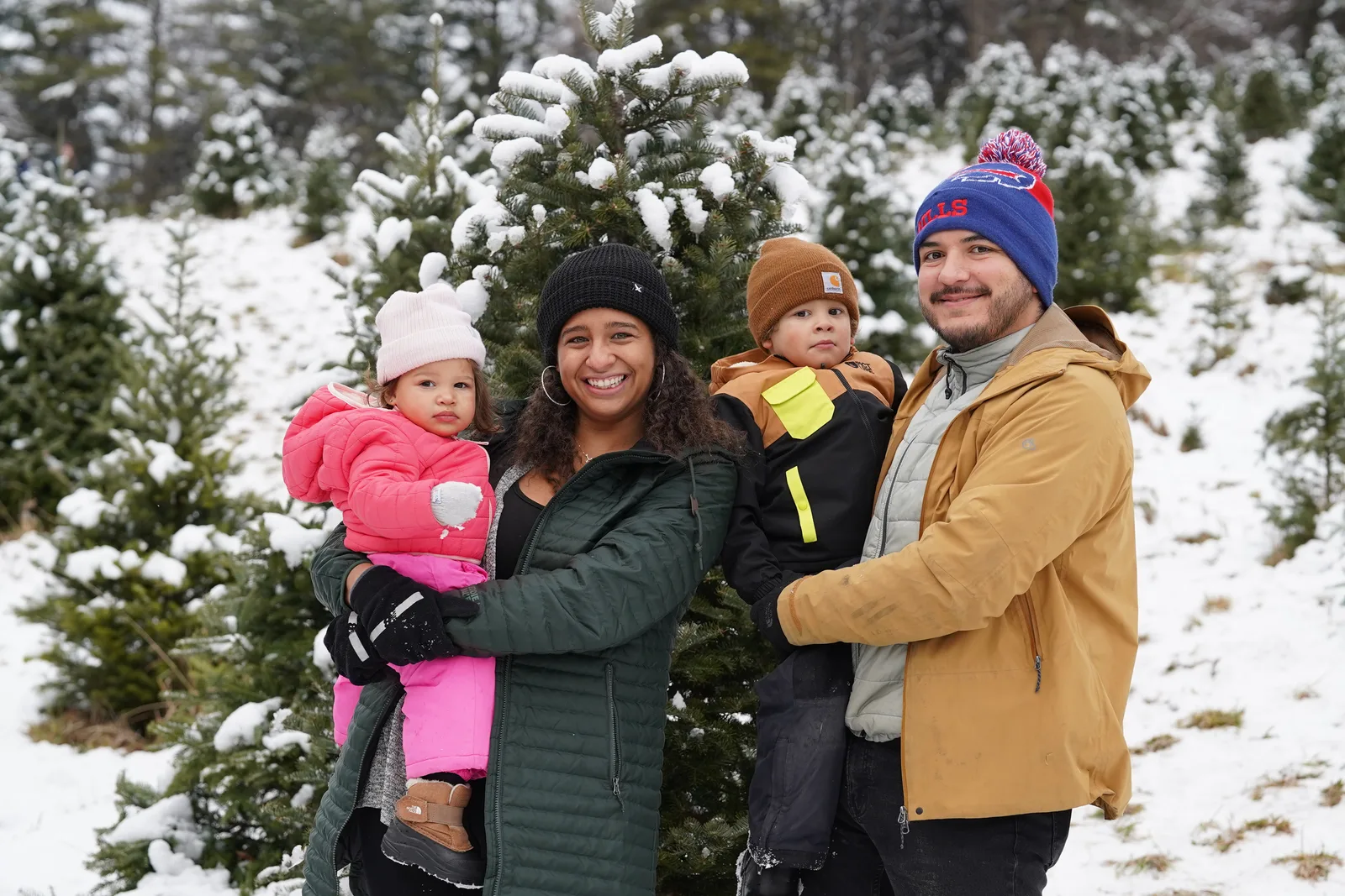 Four in winter coats pose by snow-covered evergreen, child in pink held, child in brown beanie