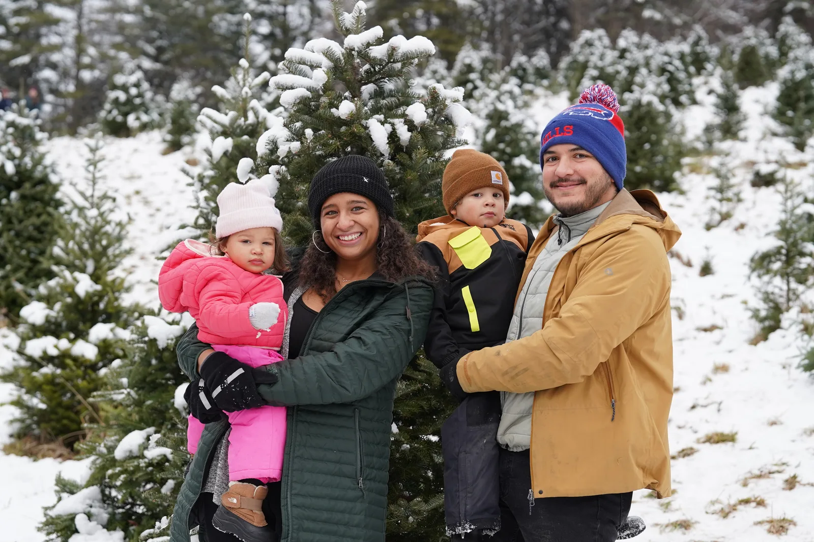 Four people in winter coats, one holding a child in pink, near snow-covered evergreen trees.
