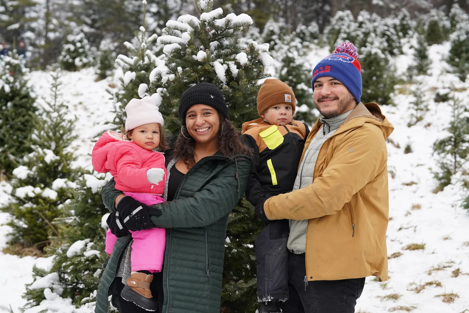 Four people in winter coats, one holding a child in pink, among snow-dusted Christmas trees
