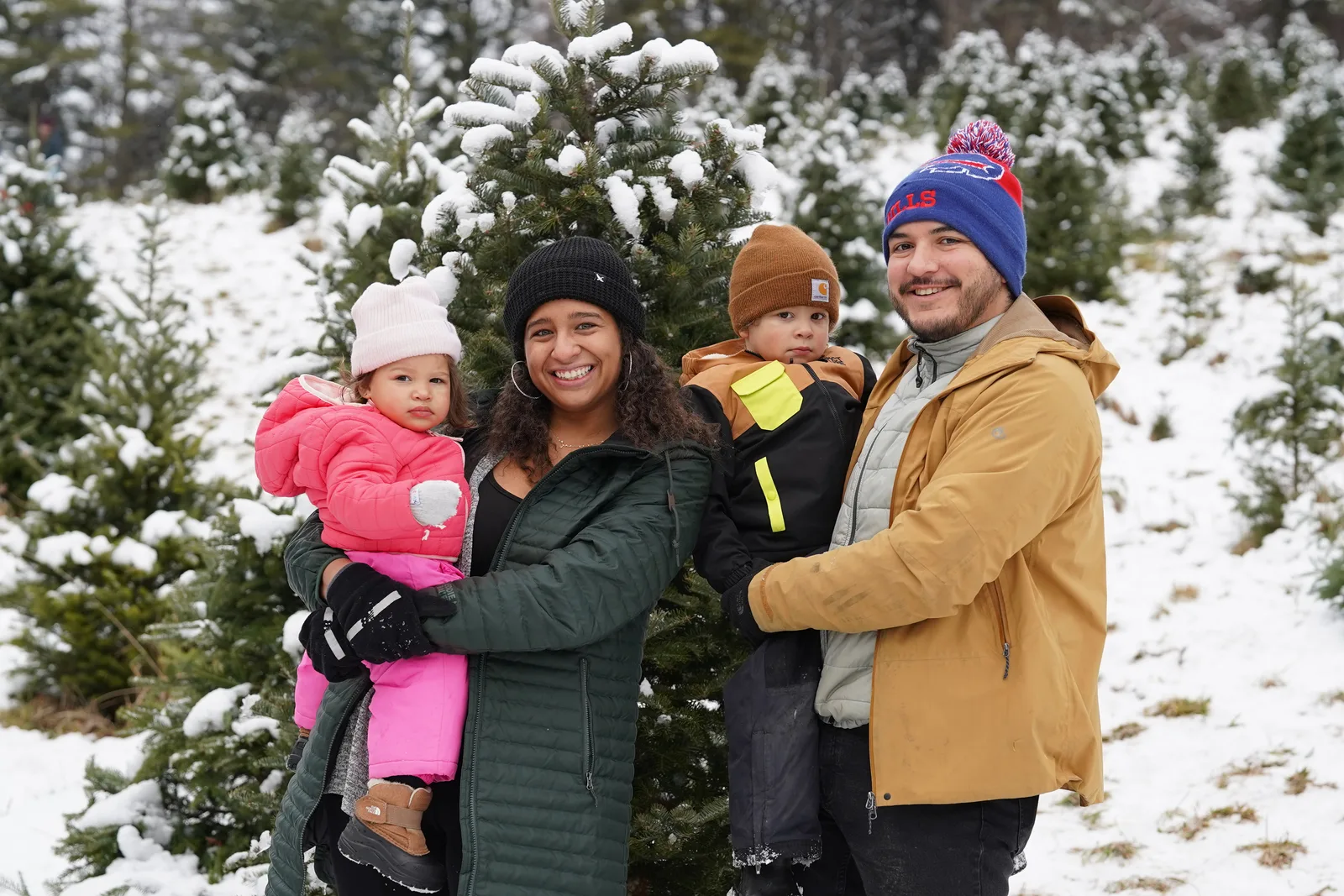 Four people in winter coats, one holding a child in pink, among snow-covered Christmas trees