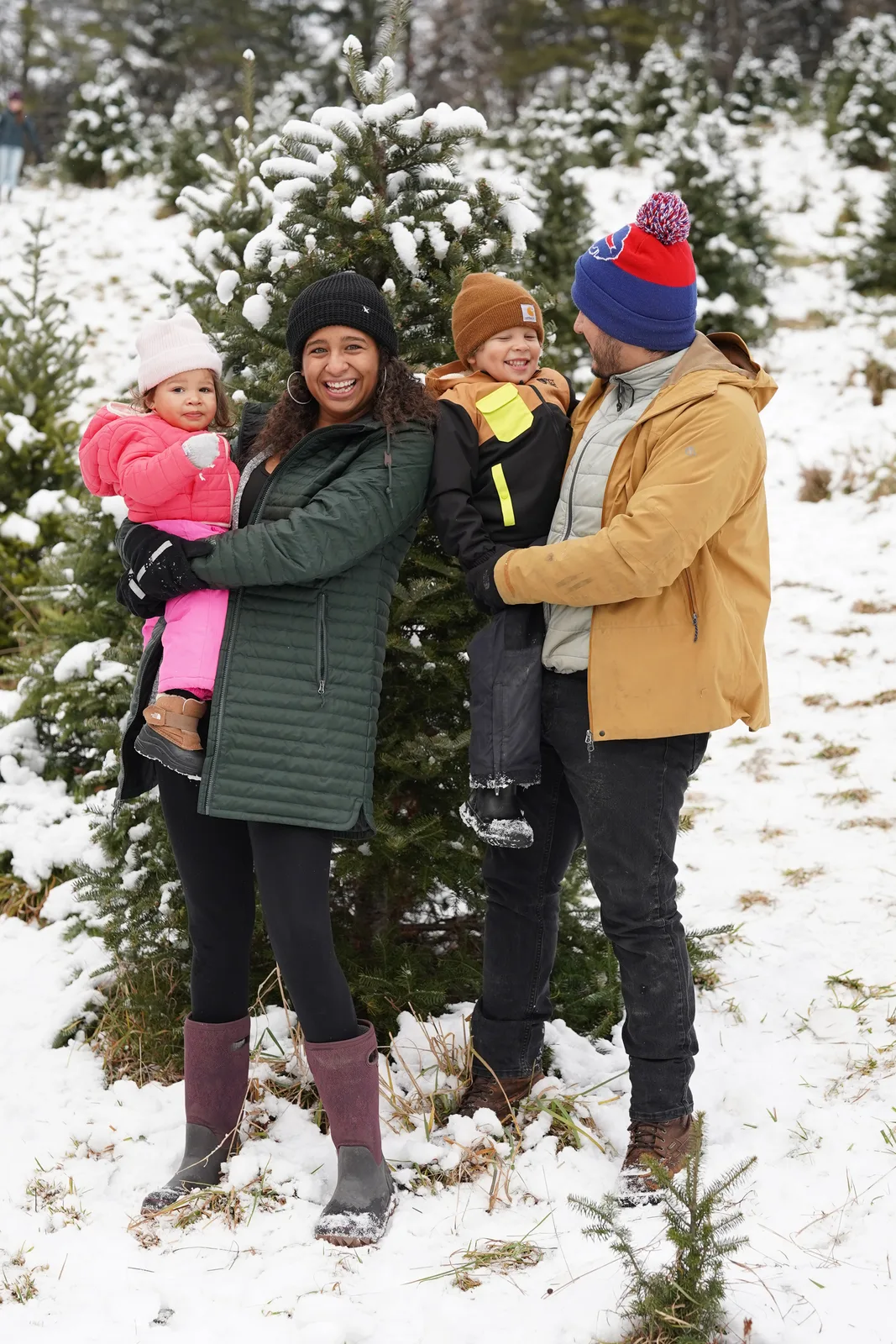 Four people in winter coats, one holding a child in pink, near snow-laden evergreen trees