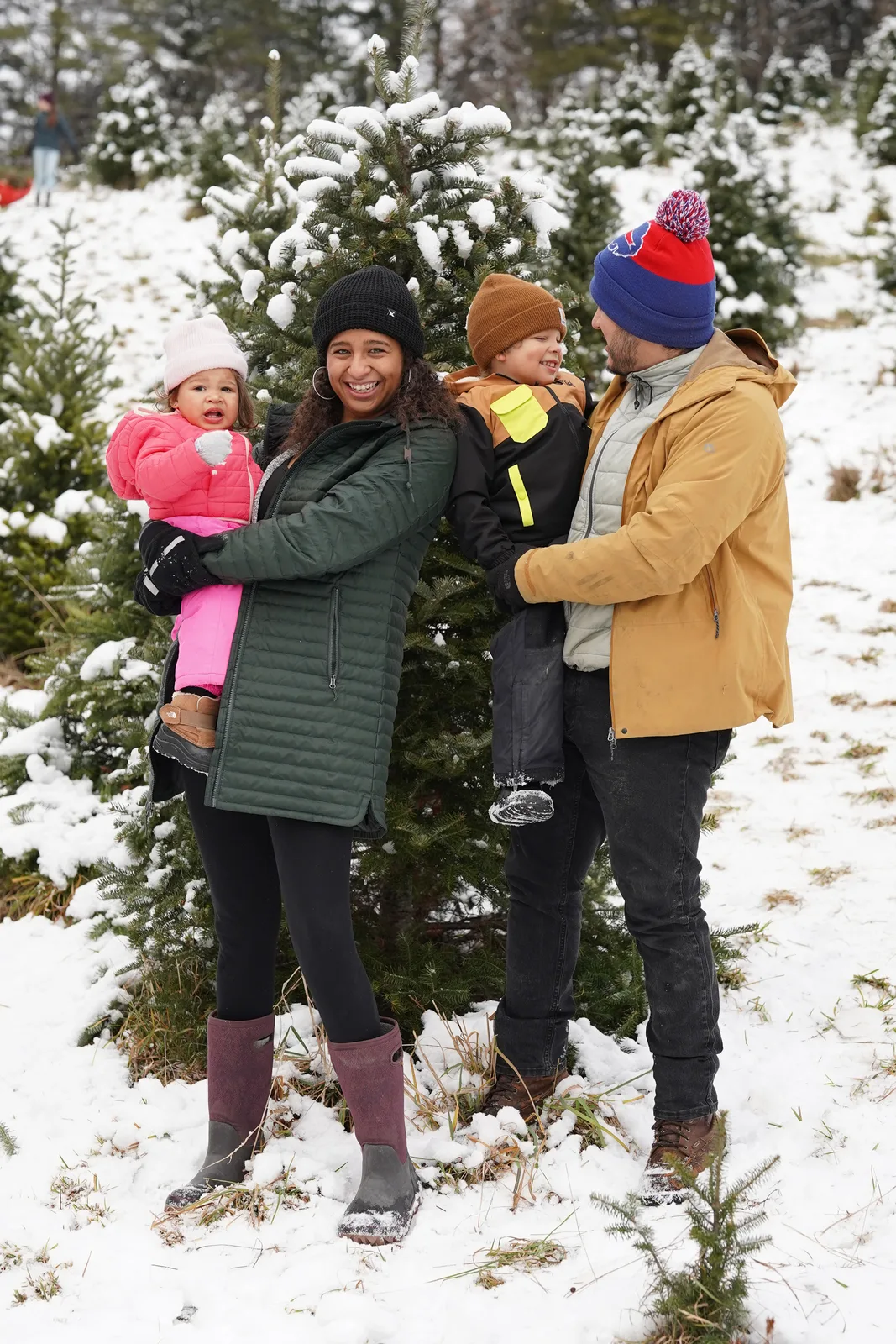 Four people in winter coats, one holding a child in pink, by snow-covered Christmas trees