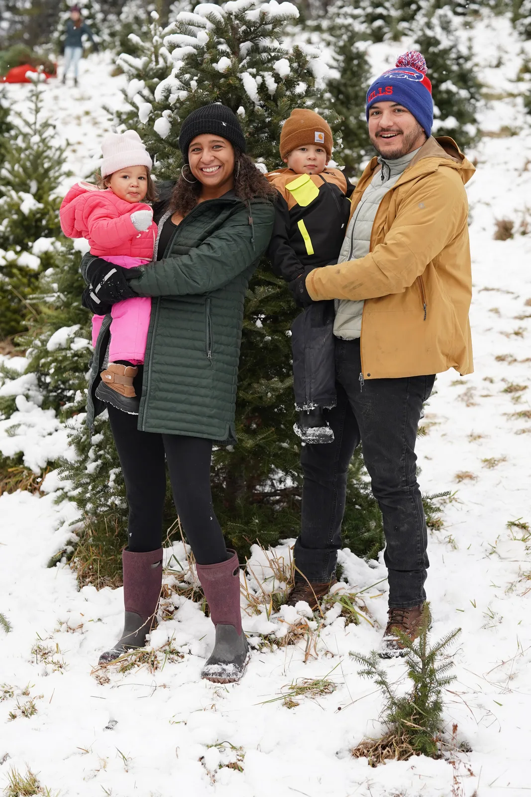 Four people in winter coats, one holding a child in pink, among snow-dusted evergreen trees