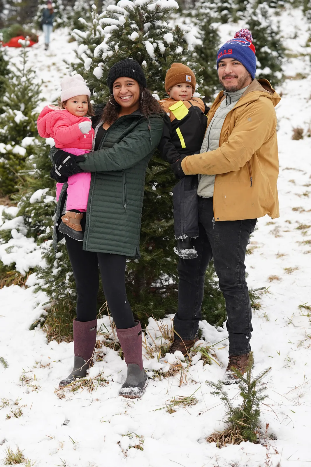 Four people in winter coats, one holding a child in pink, near snow-covered Christmas trees