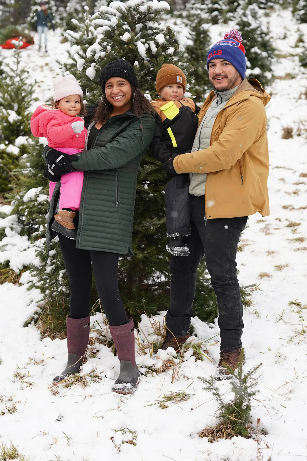 Four people in winter coats, one holding a child in pink, by snow-laden evergreen trees