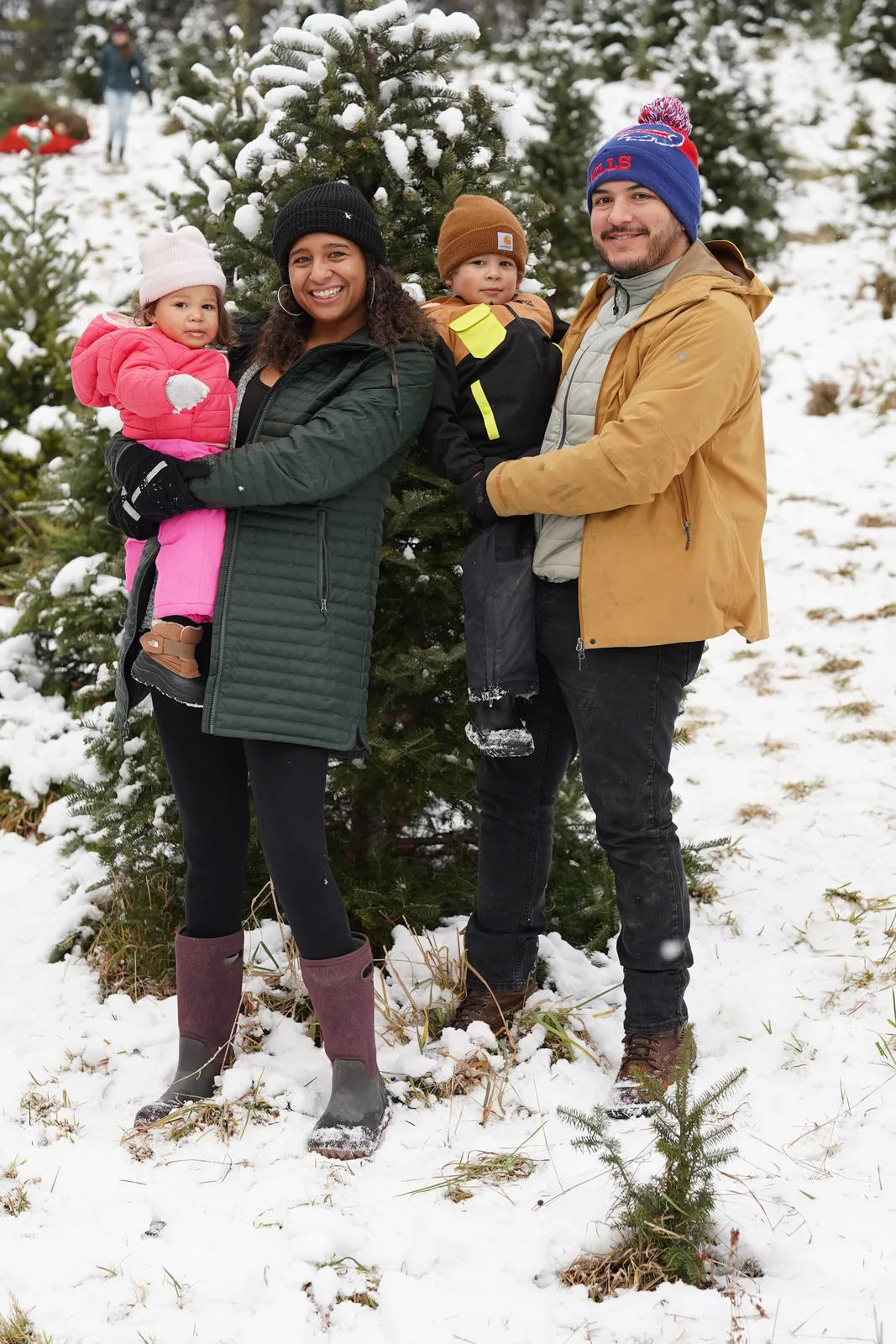 Four people in winter coats, one holding a child in pink, among snow-dusted Christmas trees