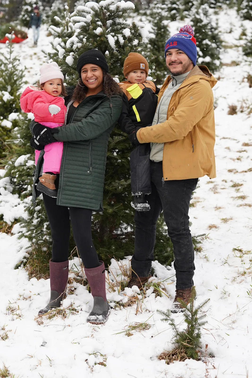 Four people in winter coats, one holding a child in pink, near snow-covered evergreen trees.