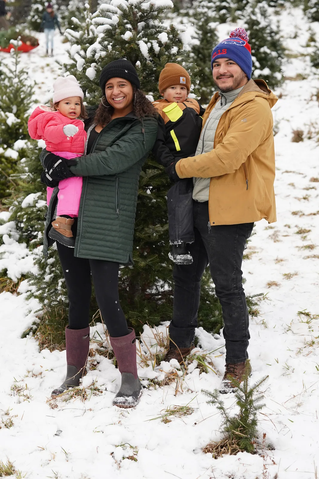 Four people in winter coats, one holding a child in pink, by snow-laden evergreen trees.