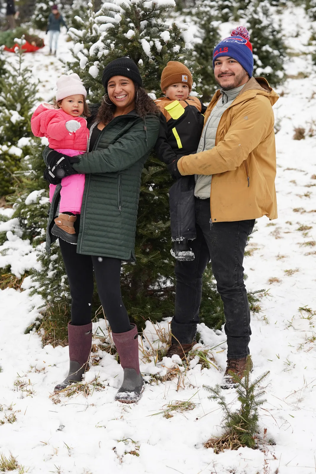Four in winter coats pose by snow-dusted evergreen, child in pink held, child in brown beanie