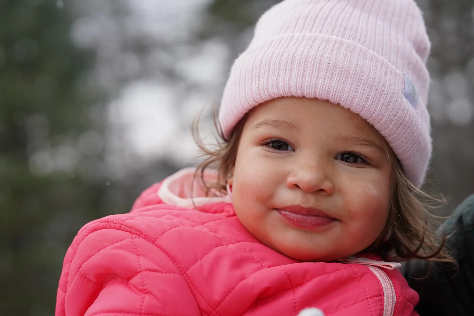 Child in pink beanie and pink quilted jacket smiling amid snow-dusted Christmas trees.