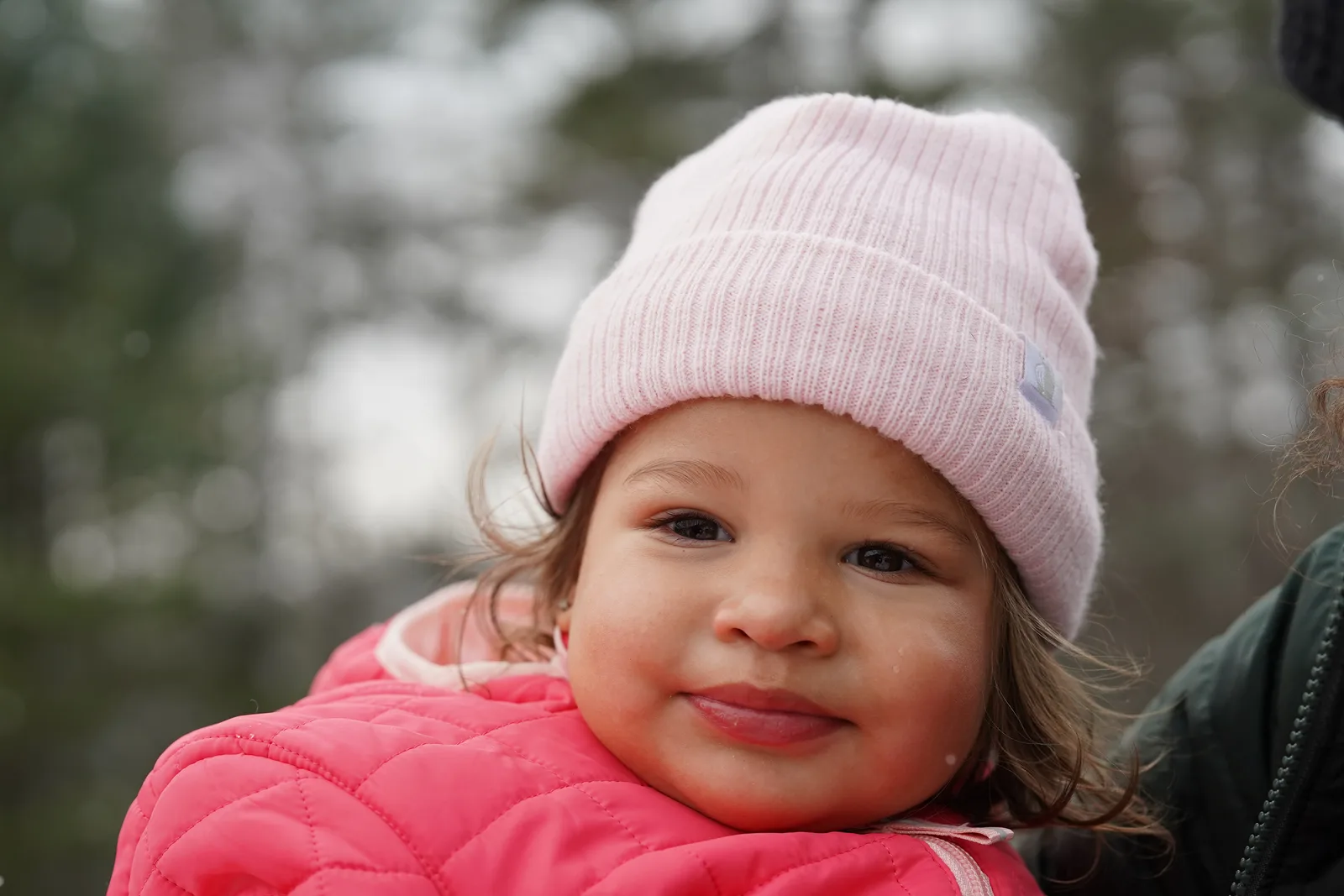 Child in pink beanie and pink quilted jacket smiling near snow-covered evergreen trees.