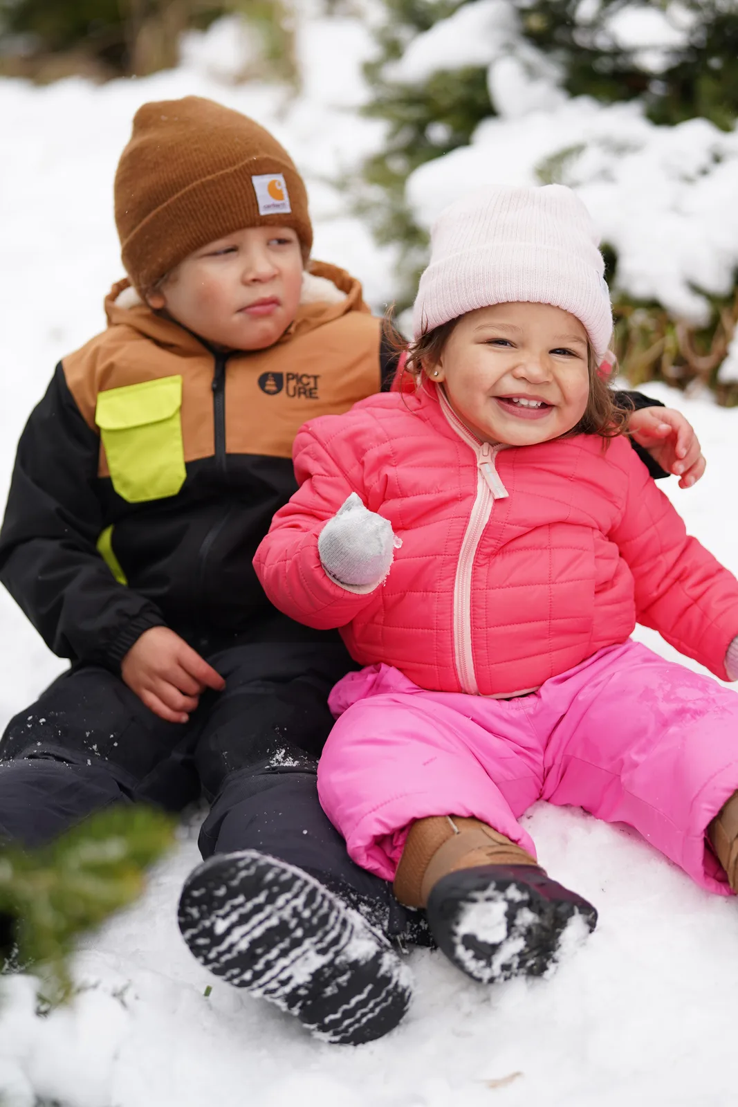 Two children in winter coats sitting in snow, one holding a snowball.