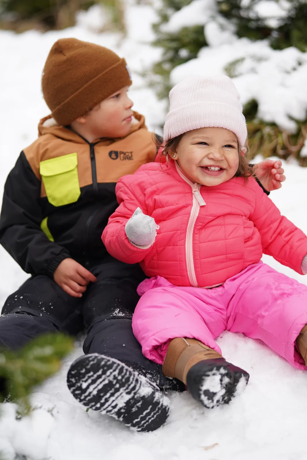 Two children in winter coats sitting in snow, one holding a snowball.