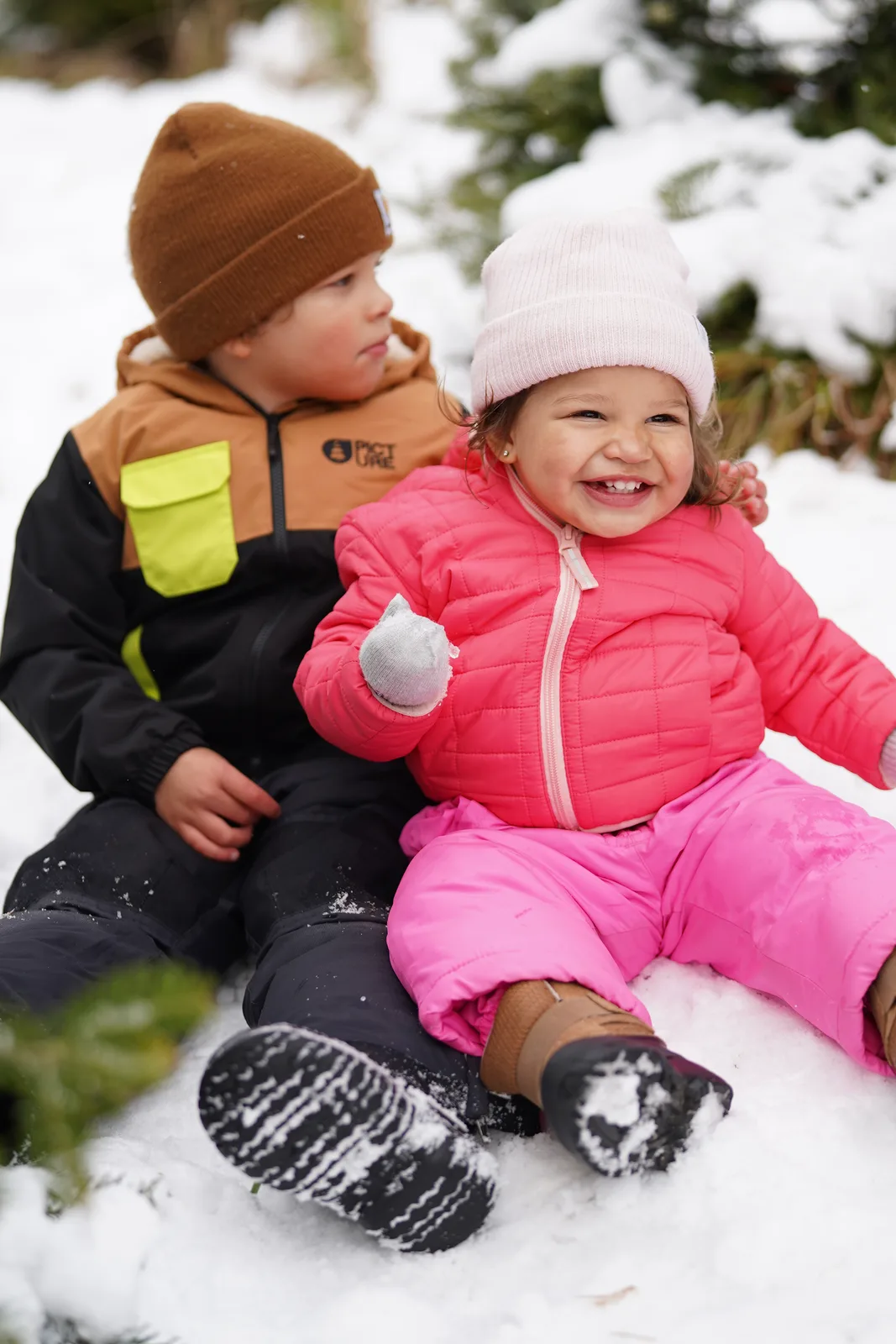 Two children in winter gear sitting in snow, one holding a snowball, evergreens behind.