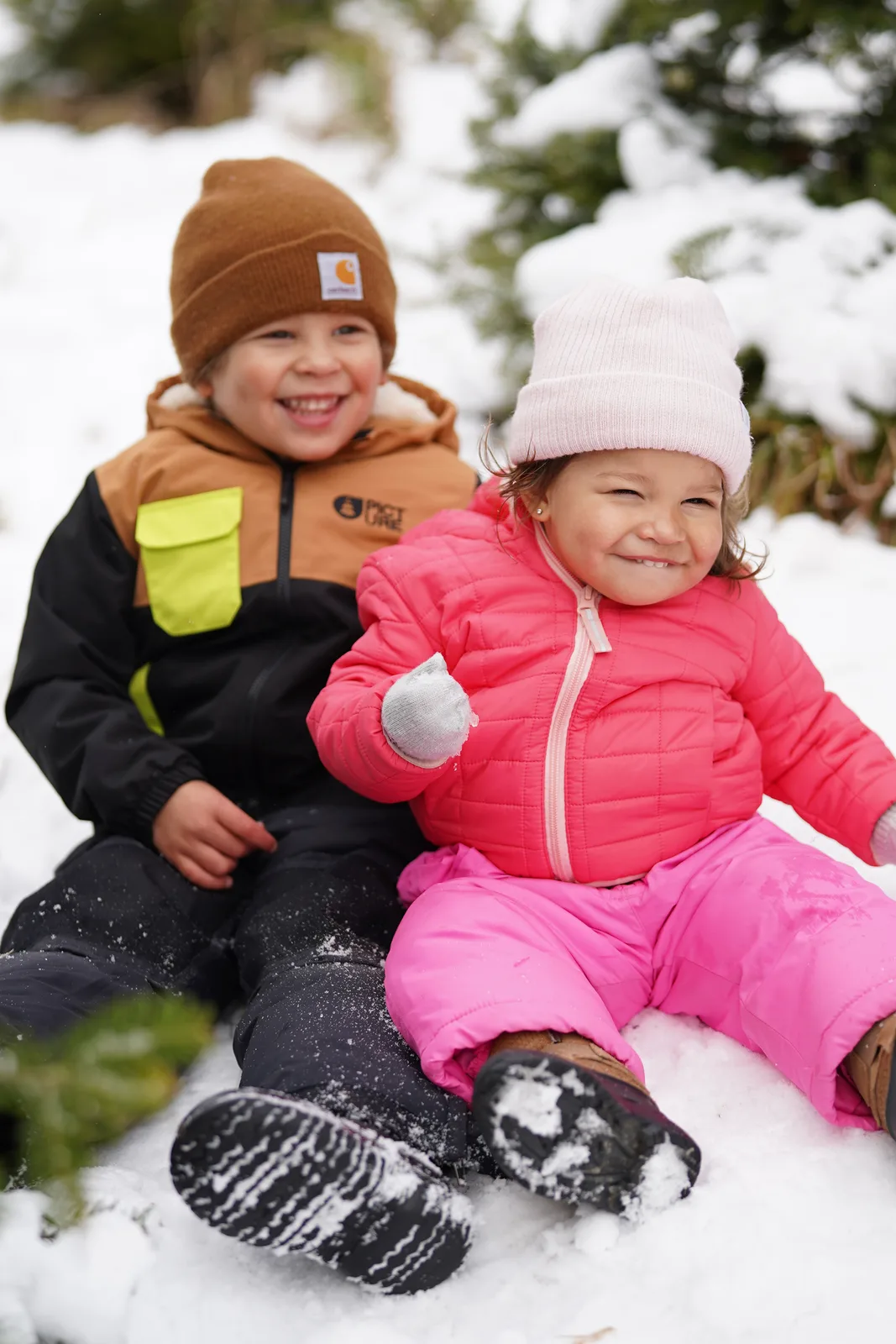Two children in winter clothing sitting in snow, one holding a snowball.