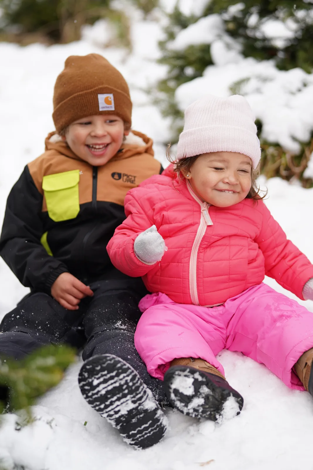 Two children in winter coats and hats sitting in snow, snow-covered trees behind.