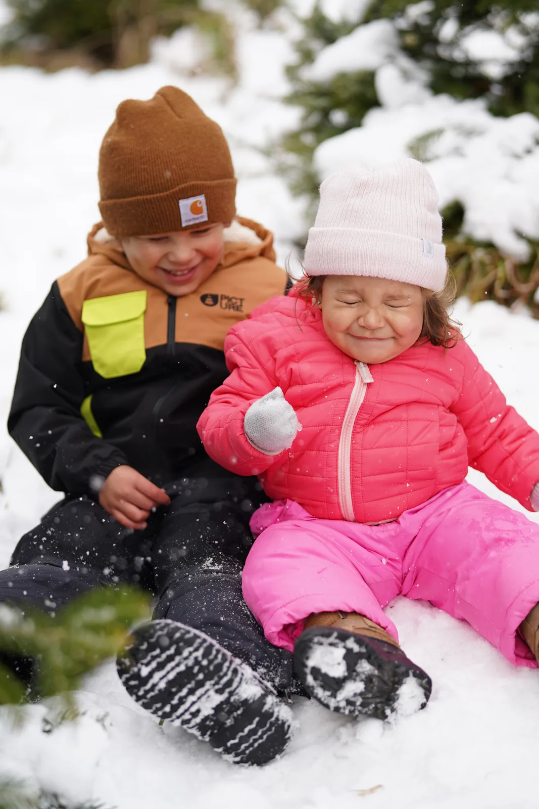 Two children in winter clothing sitting in snow, one holding a snowball, snow falling.