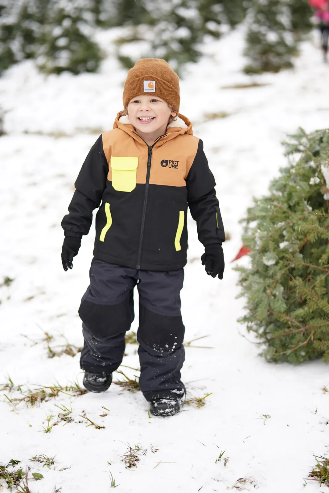 Child in brown beanie and black jacket with yellow accents standing near snow-dusted evergreen.