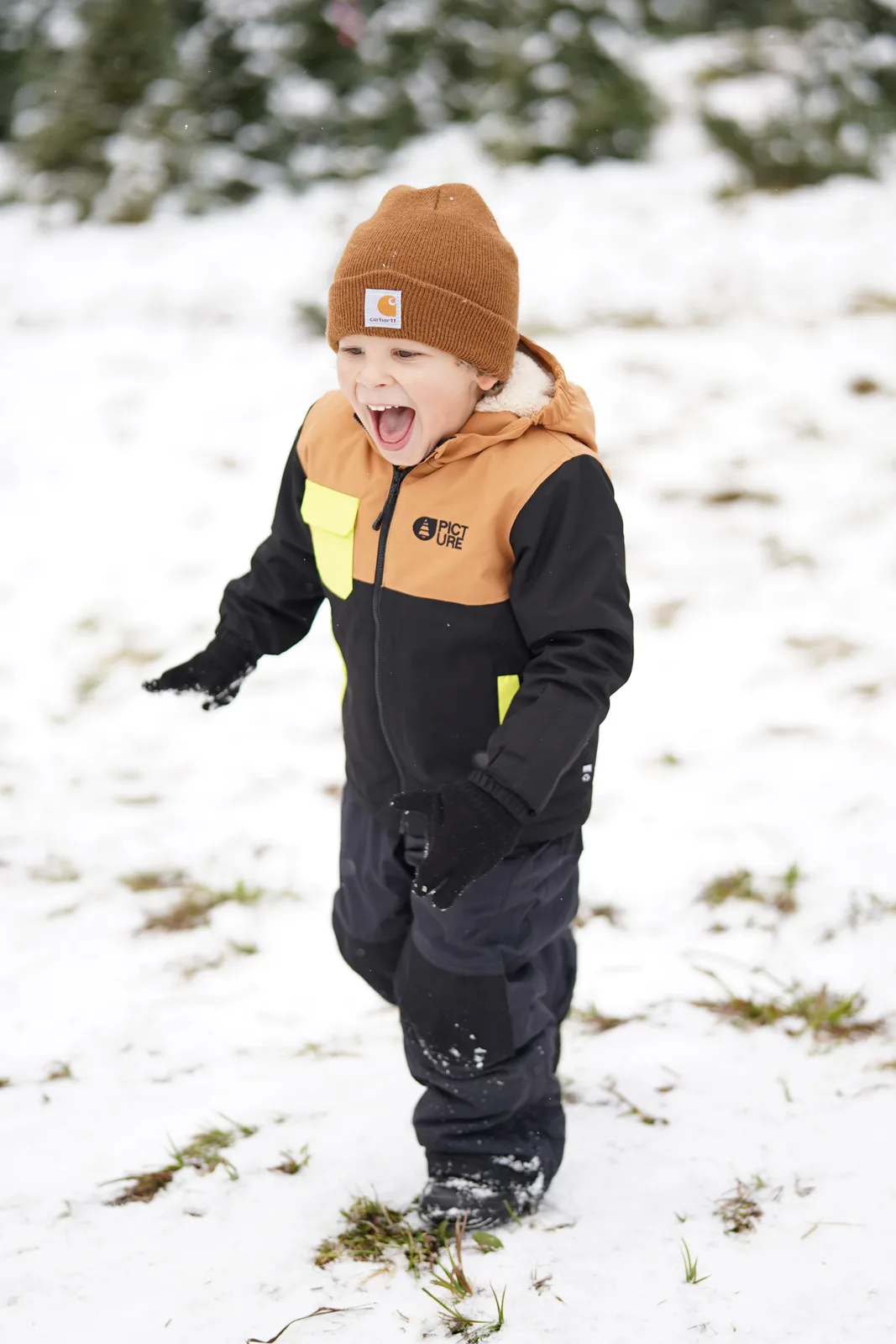 Child in brown beanie and black jacket with yellow accents shouting in snowy field.