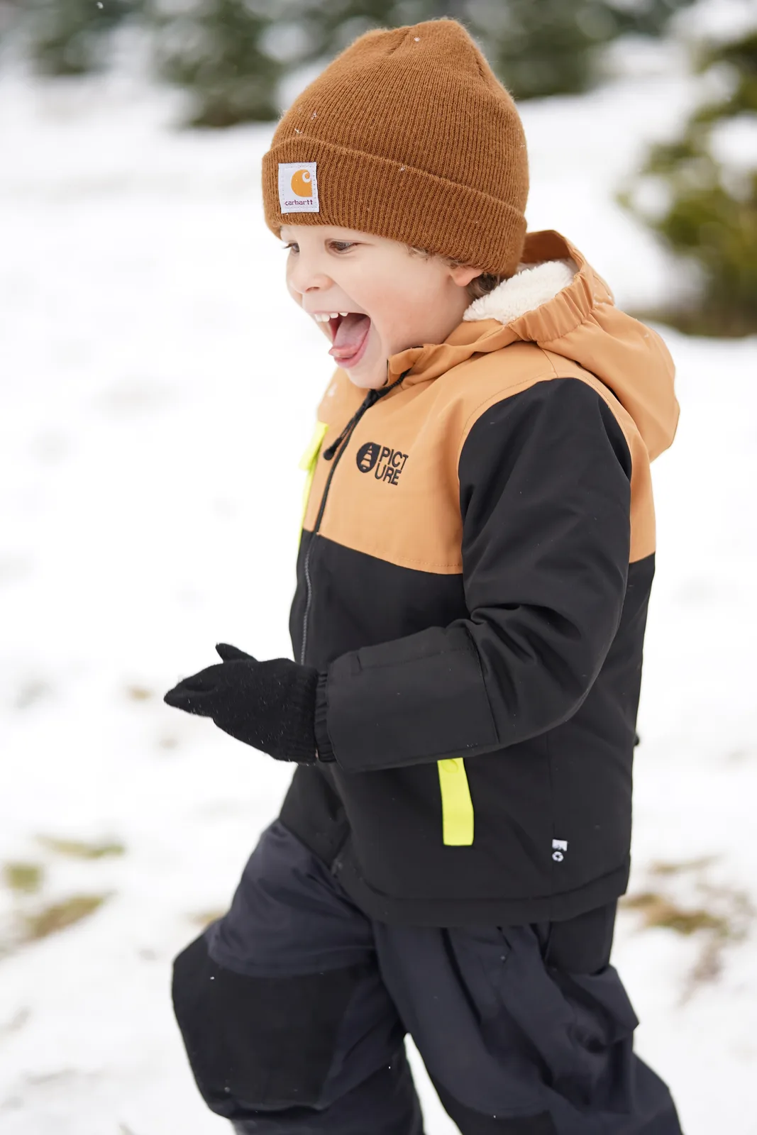 Child in brown beanie and black jacket with yellow accents laughing in snow.