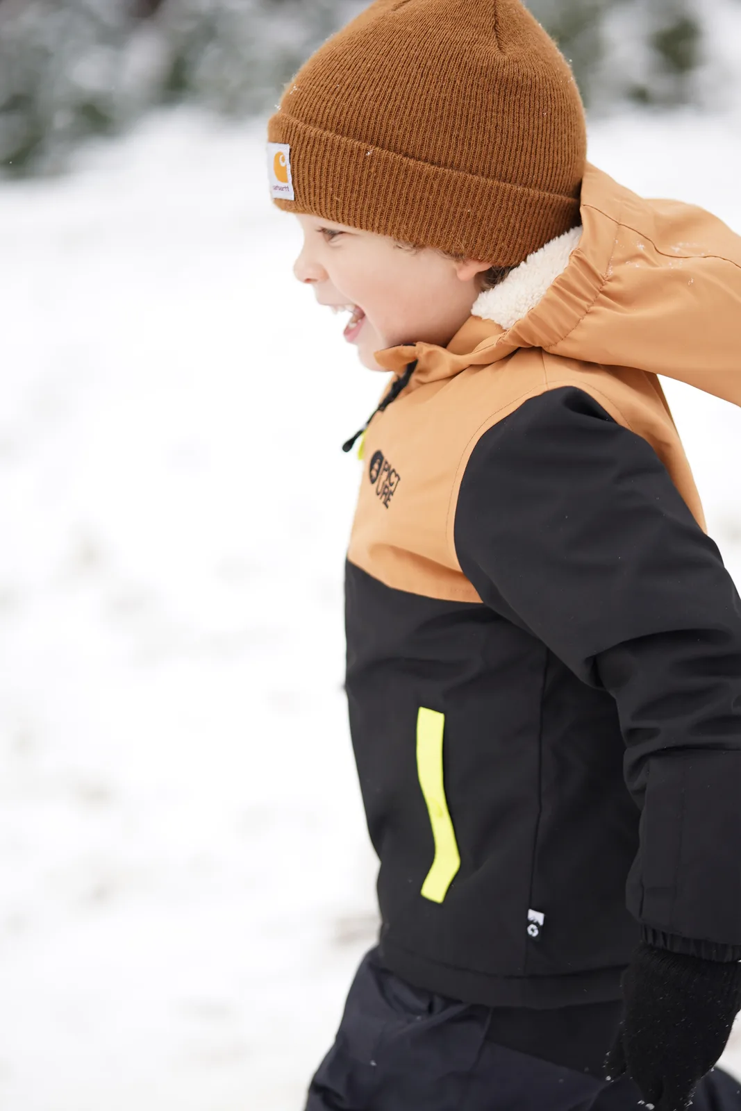 Child in brown beanie and black jacket with yellow accents looking up in snow.