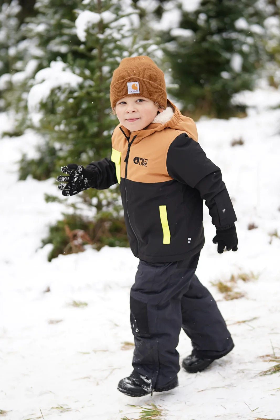 Child in brown beanie and black jacket with yellow accents holding snowball in snow.