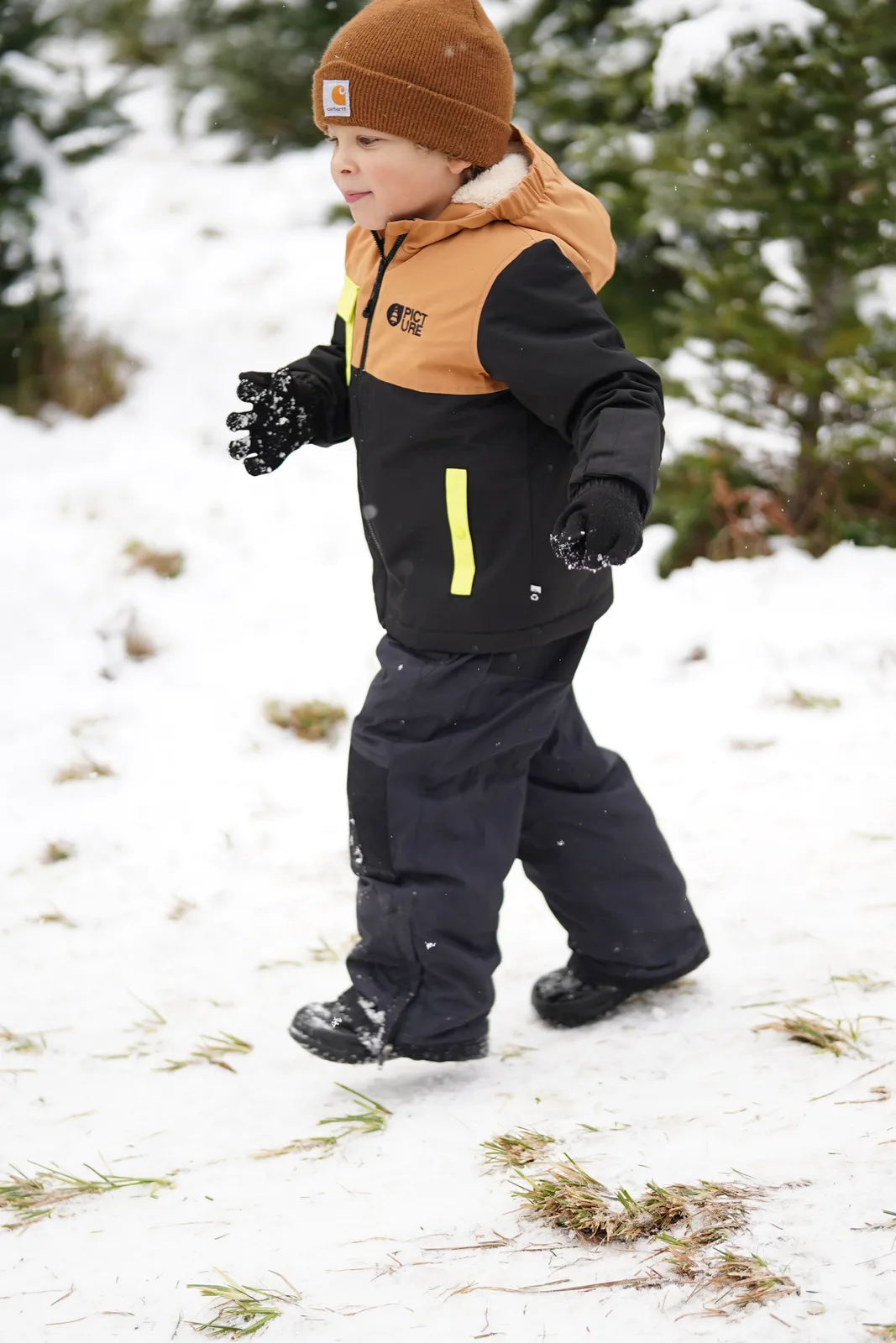 Child in brown beanie and black jacket with yellow accents walking in snow.