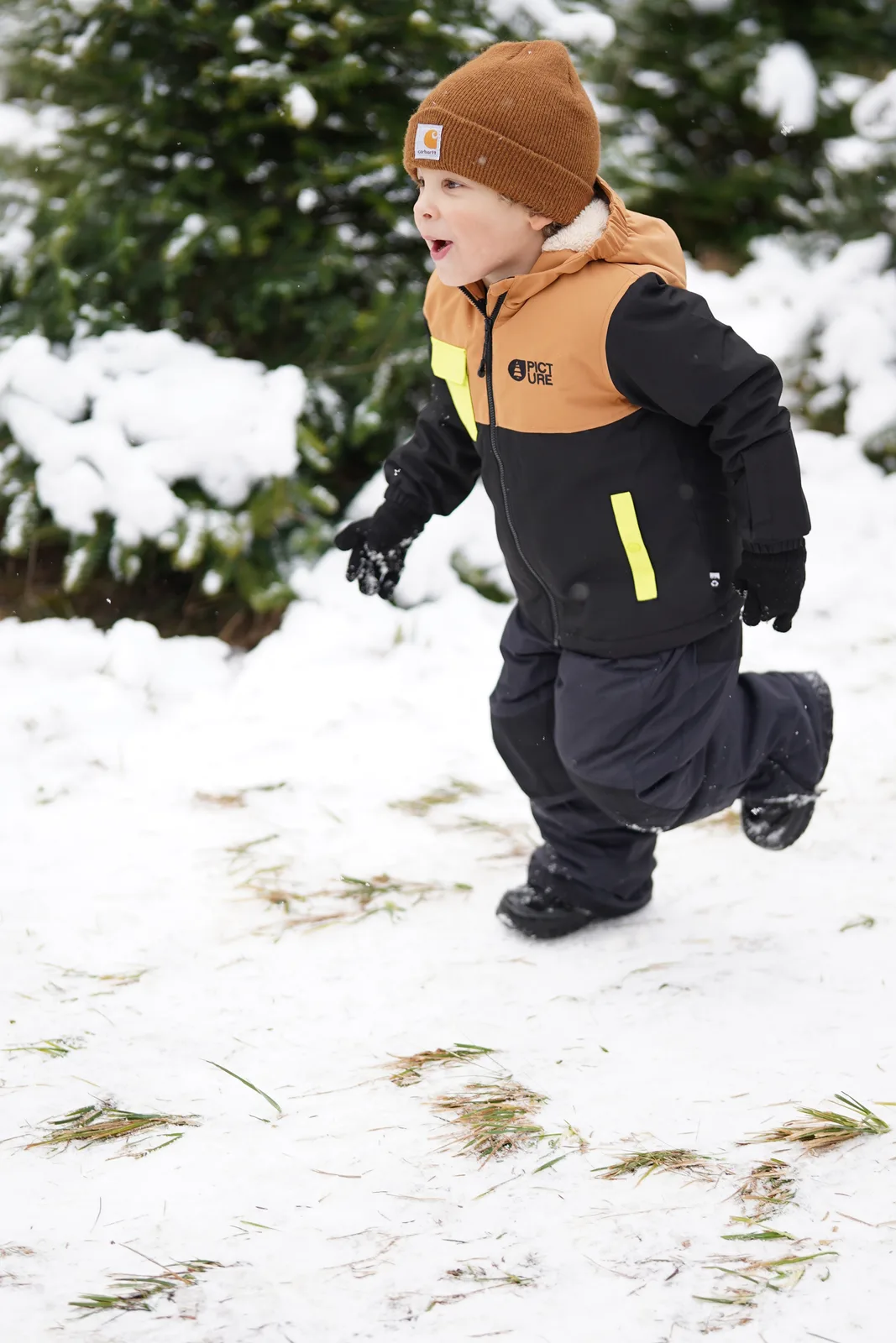 Child in brown beanie and black jacket with yellow accents running in snow.