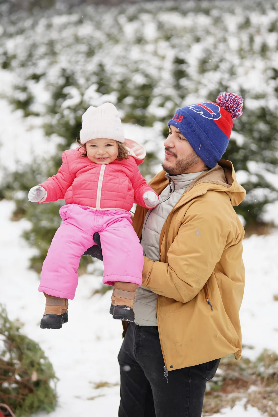 Adult in tan jacket and blue beanie holding child in pink snowsuit, snow-dusted evergreens nearby