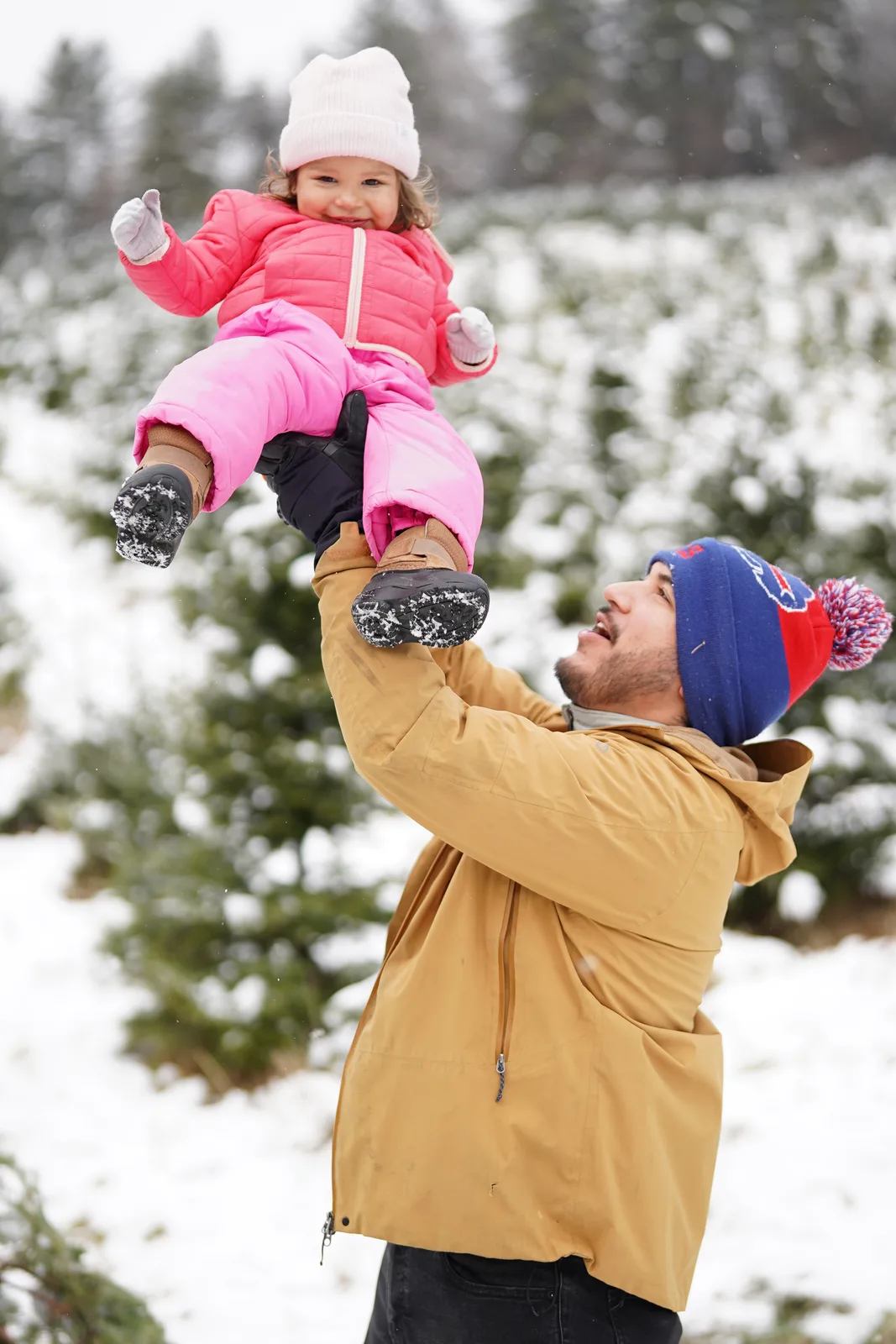 Adult in tan jacket and blue beanie lifting child in pink snowsuit, snow falling among trees