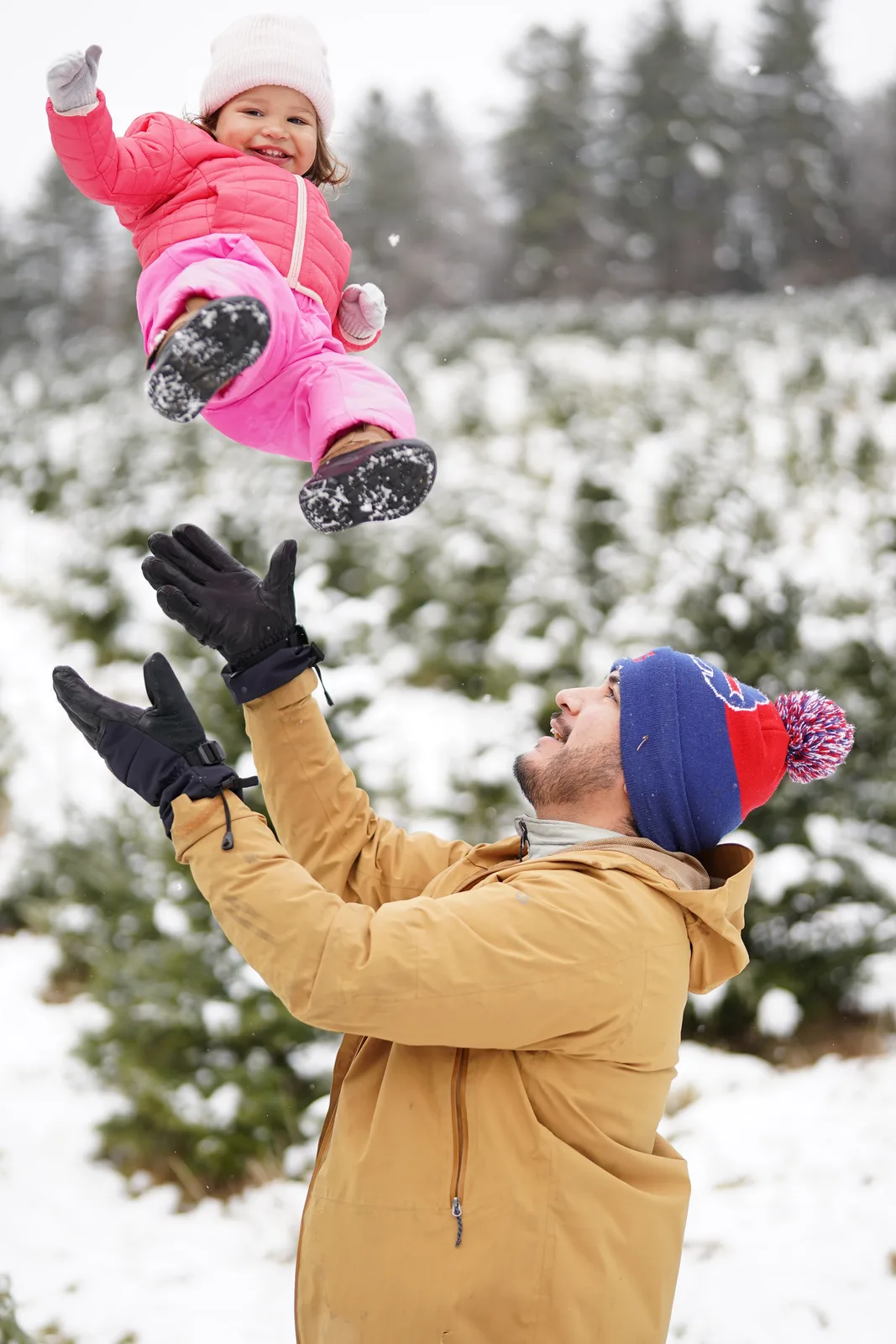 Adult in tan jacket and blue beanie tossing child in pink snowsuit, snow-covered trees in background