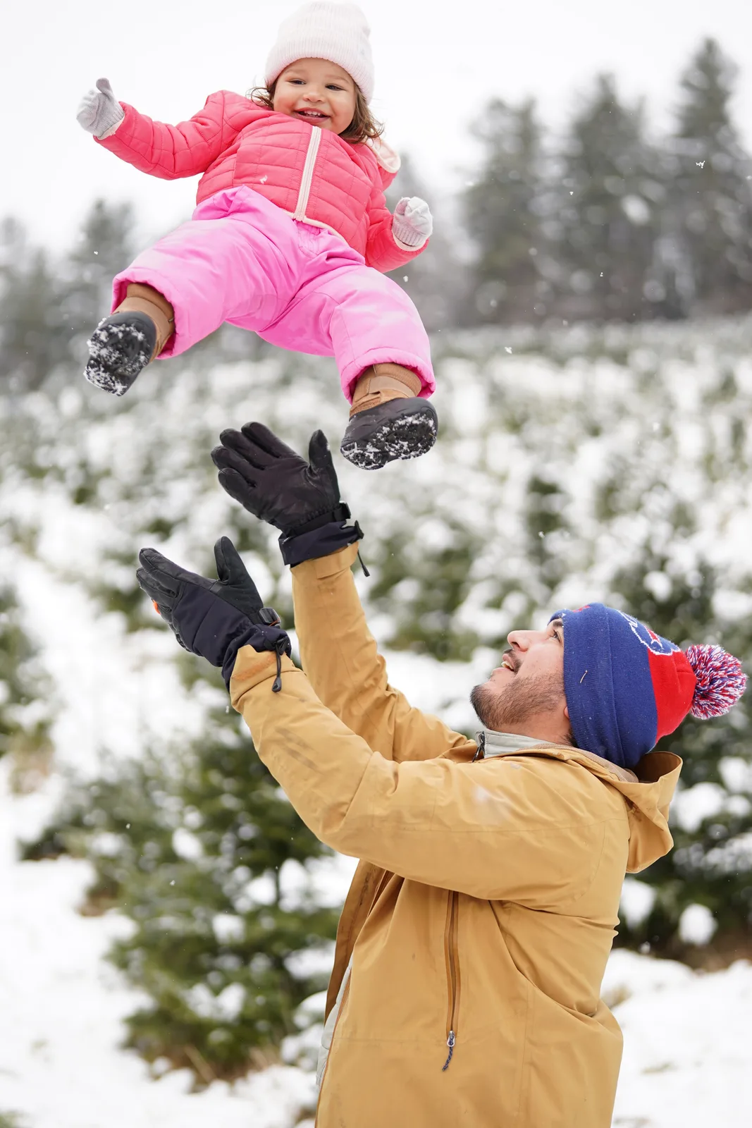 Adult in tan jacket and blue beanie catching child in pink snowsuit, snow falling on evergreens
