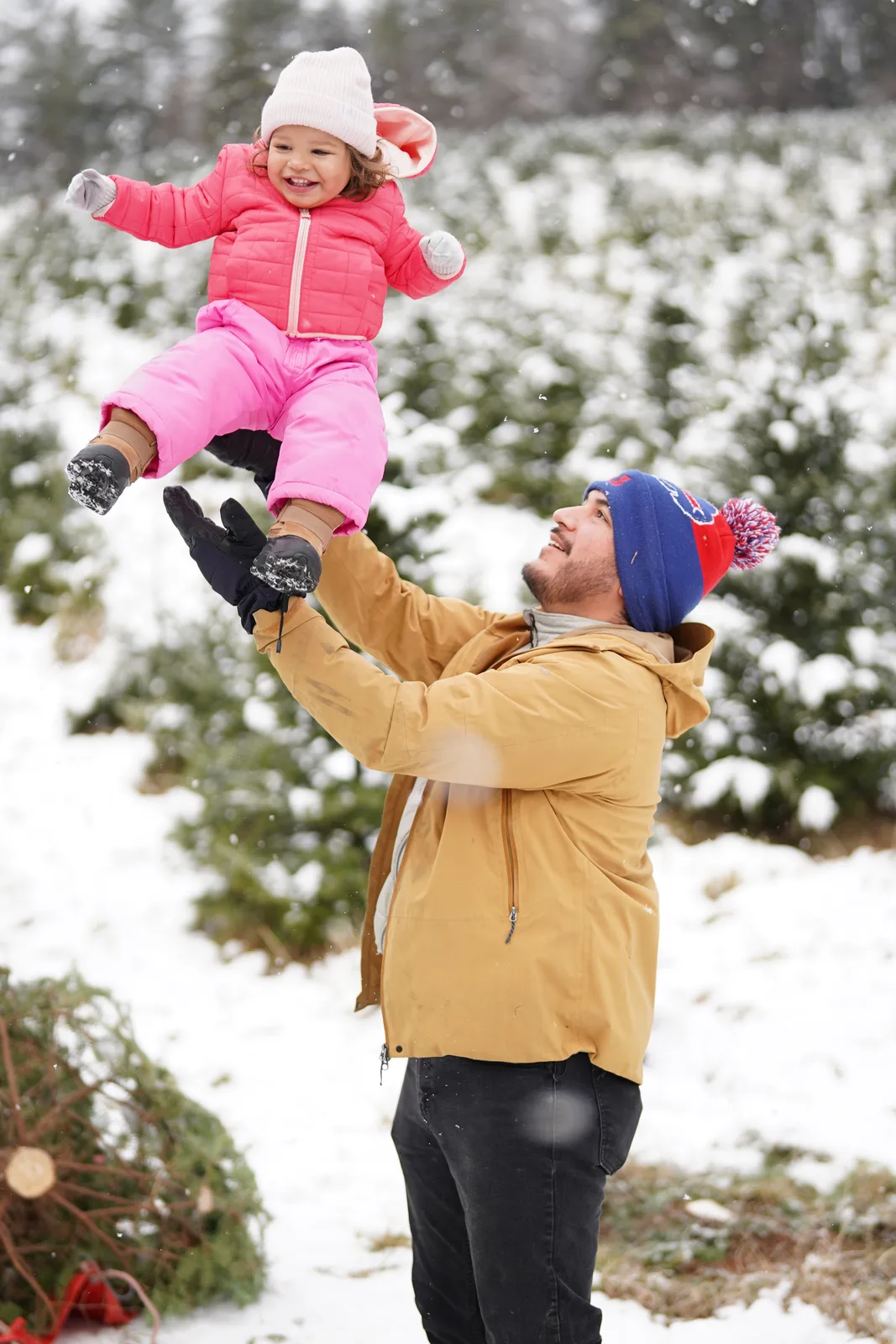 Adult in tan jacket and blue beanie holding child in pink snowsuit, snow falling among evergreens
