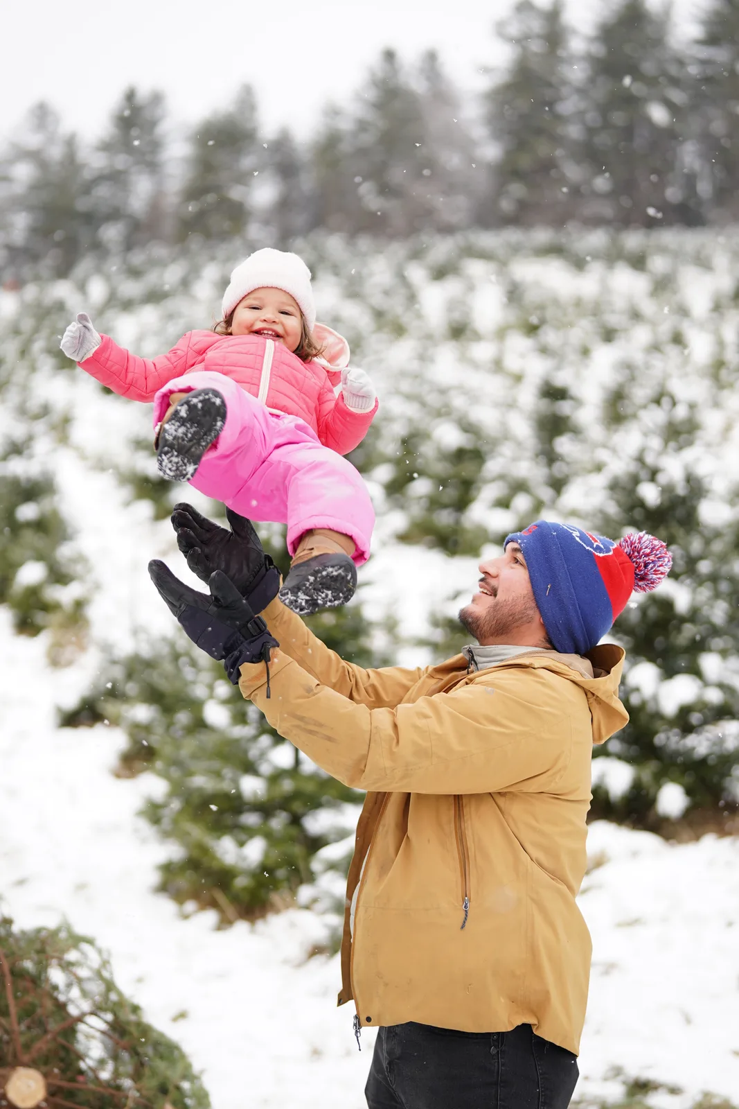 Person in tan jacket and blue beanie lifting child in pink snowsuit among snow-covered trees