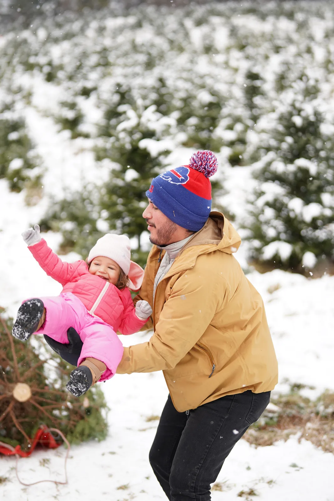 Person in tan jacket and blue beanie holding child in pink snowsuit near snow-laden evergreens