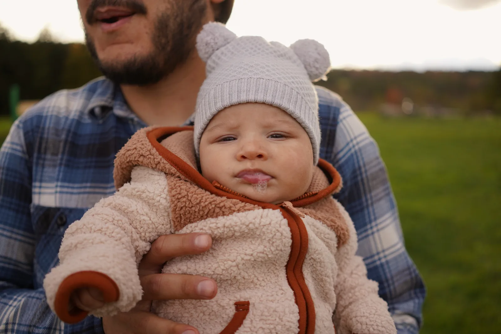 Adult holding a baby in a light gray bear hat and cream jacket outdoors