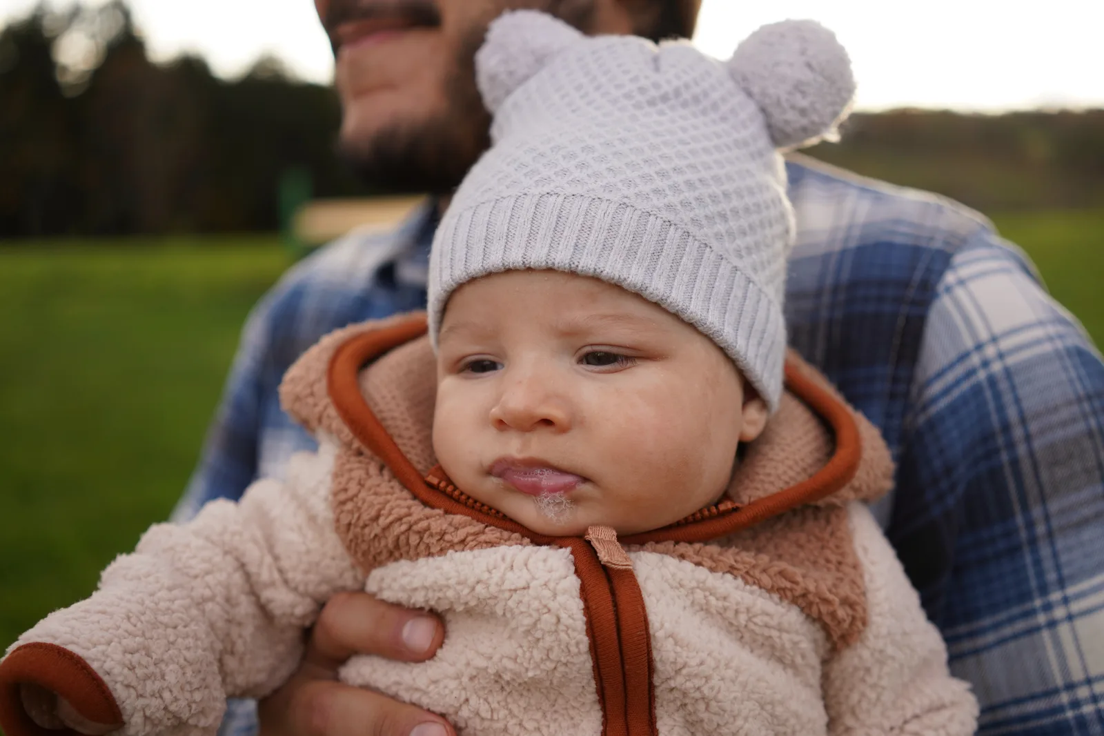 Adult holding a baby in a light gray bear hat and cream jacket in a field