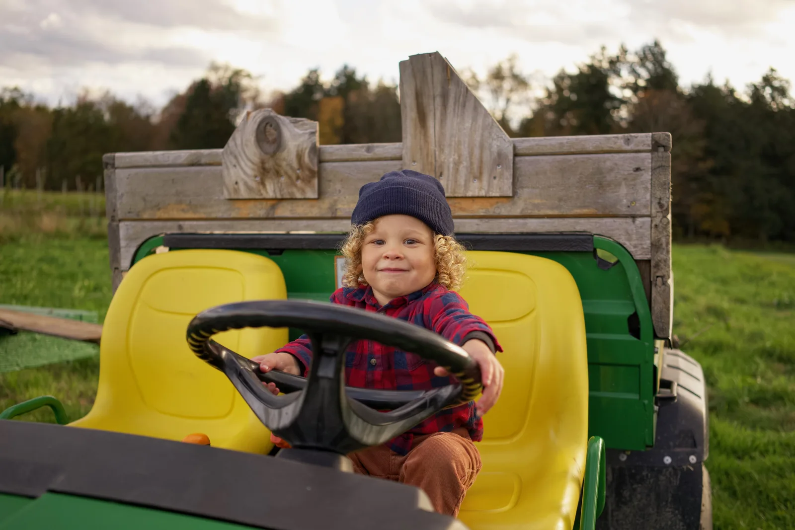 Child in a blue beanie and red plaid shirt steering a green tractor with yellow seat