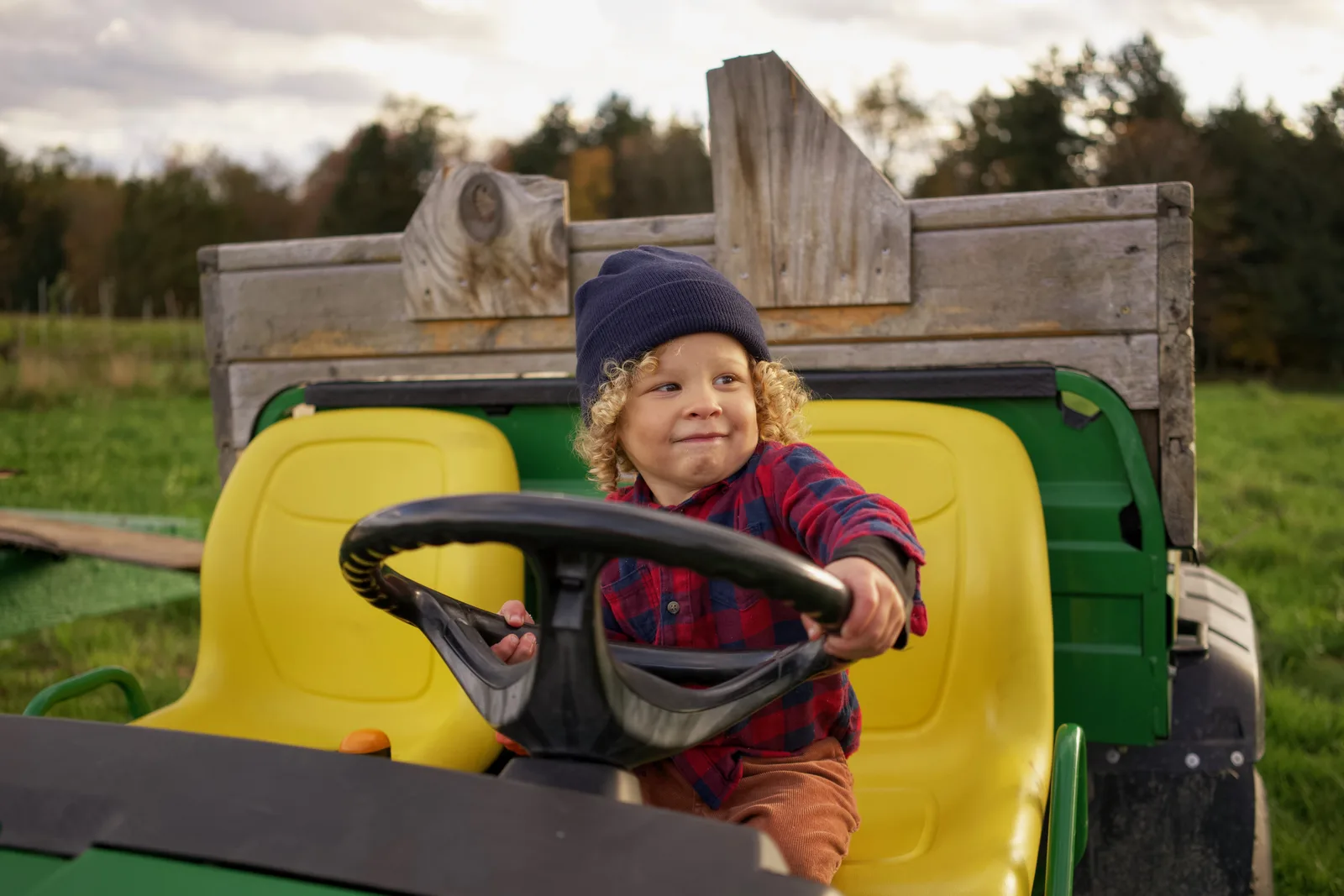 Child in a blue beanie and red plaid shirt driving a green tractor in a field