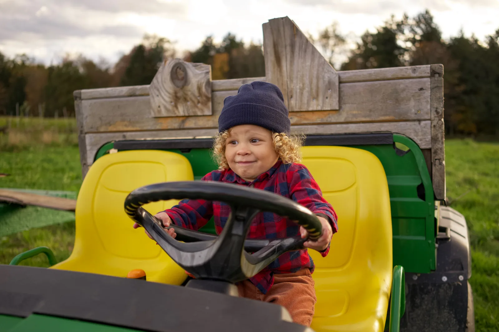 Child in a blue beanie and red plaid shirt steering a green tractor outdoors