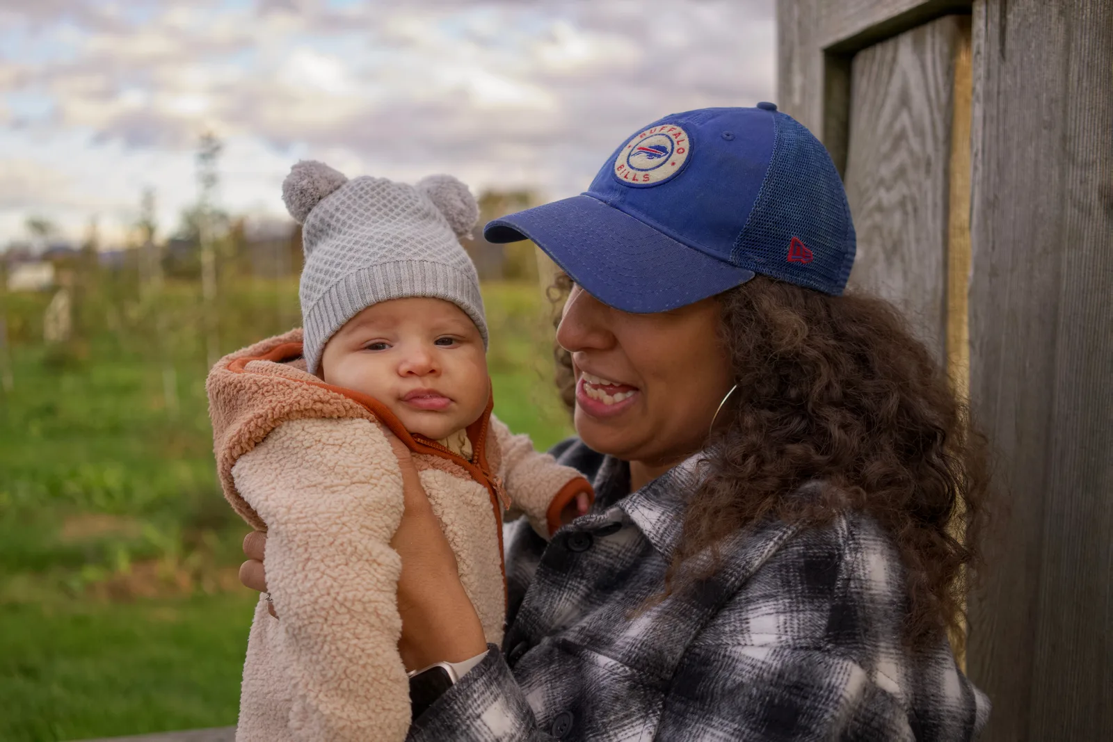 Adult holding a baby in a light gray knit hat and cream jacket by a wooden structure