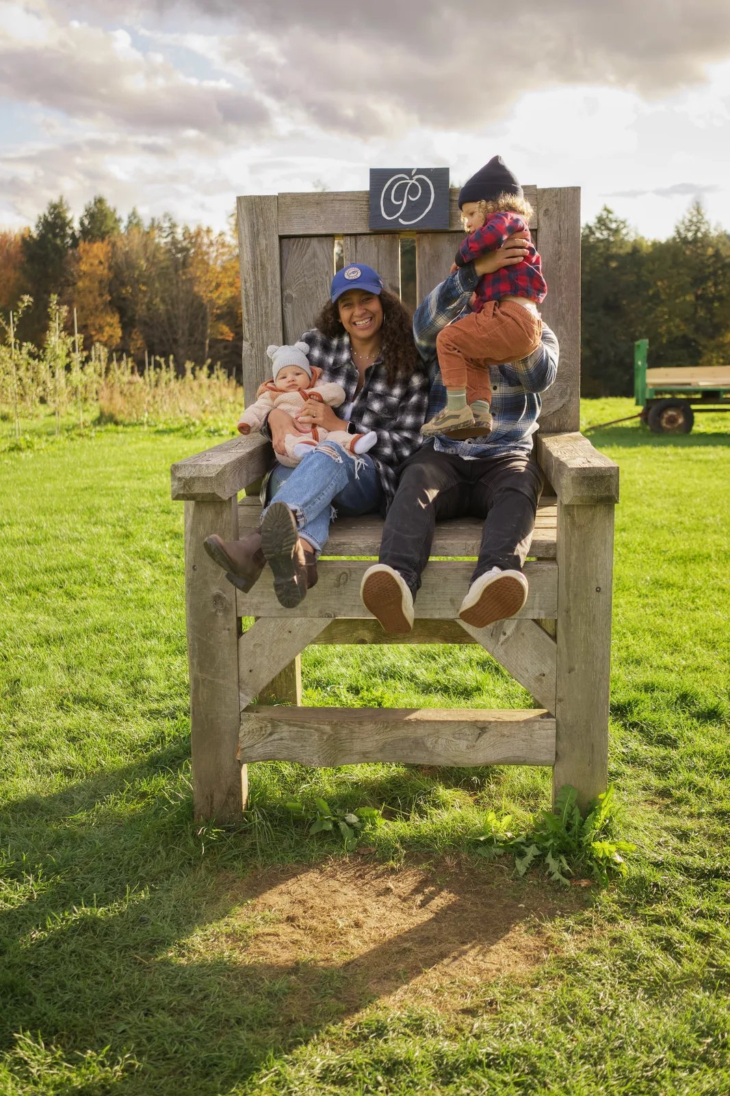 Three people on a large wooden chair in a grassy field with autumn trees