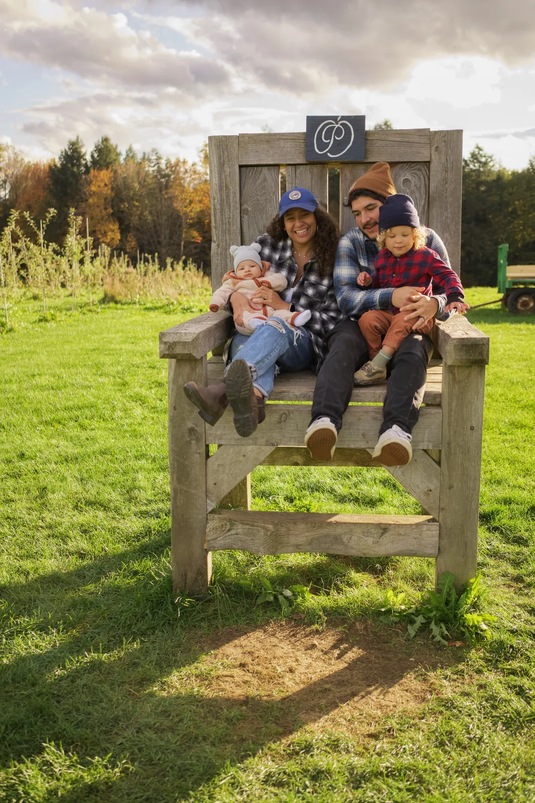 Four people on large wooden chair in grassy field, autumn trees and orchard rows behind