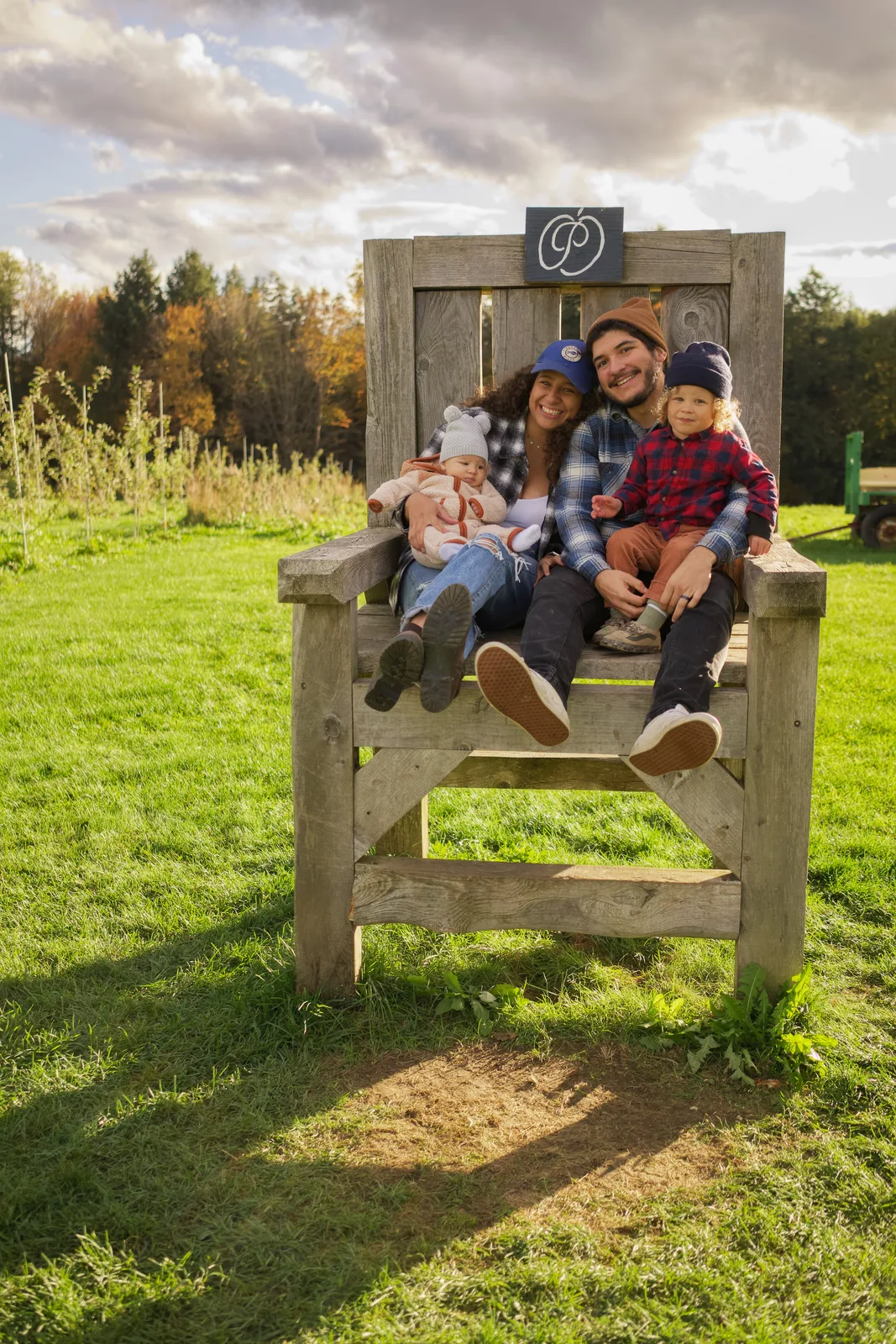Four people on wooden chair, green grass and cloudy sky over orchard