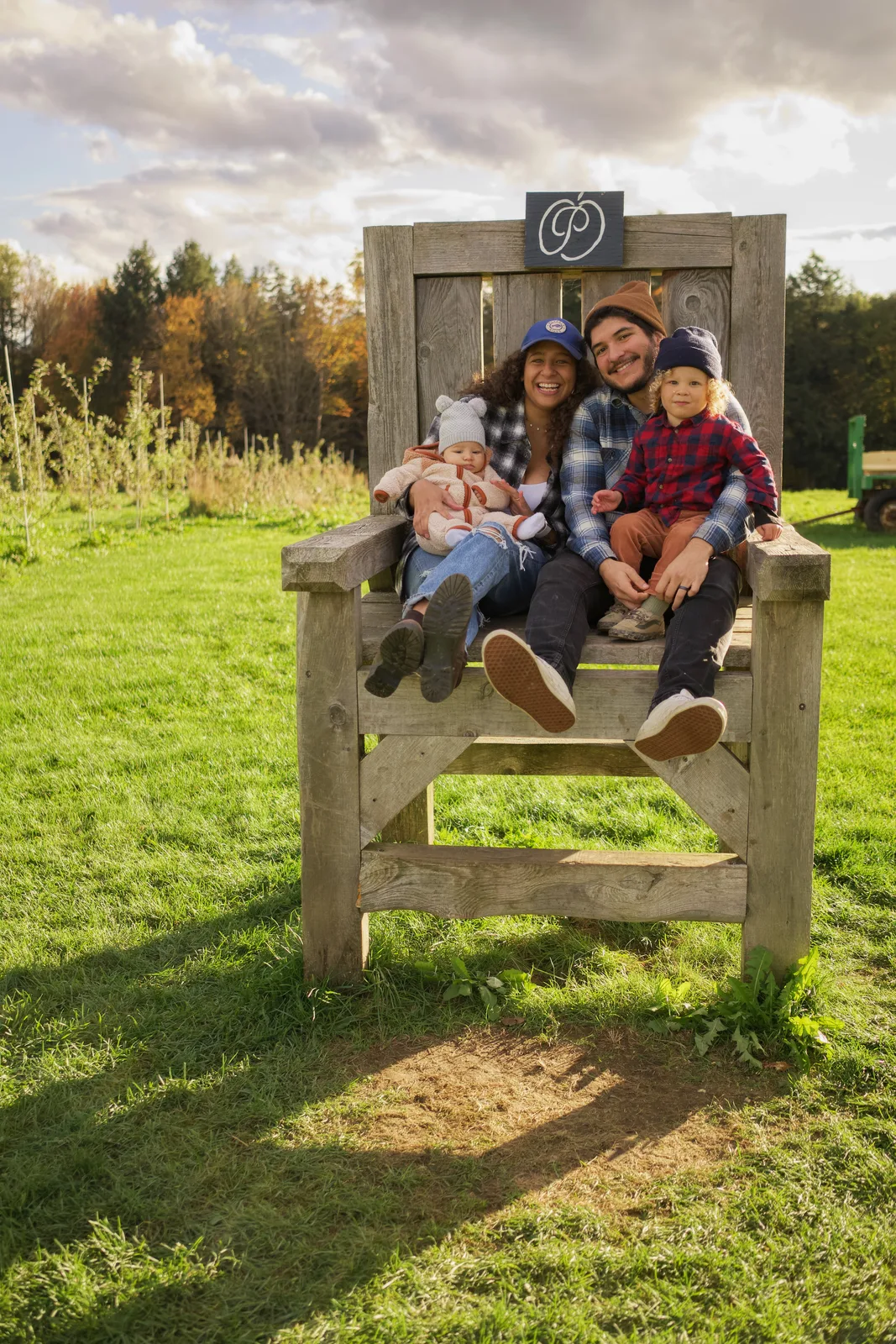 Four people on large wooden chair, grassy field and orchard rows with autumn foliage behind