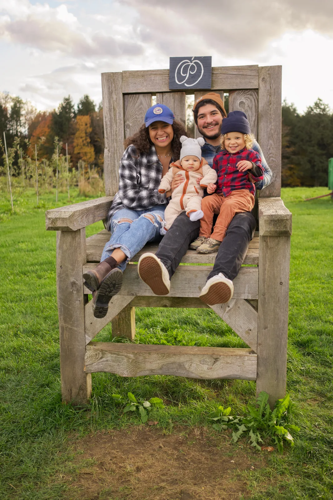 Four people on large wooden chair with sign above, grassy field and orchard rows behind