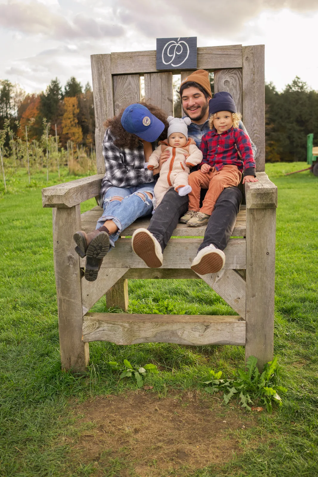 Four people seated on a large wooden chair in a grassy field with autumn trees