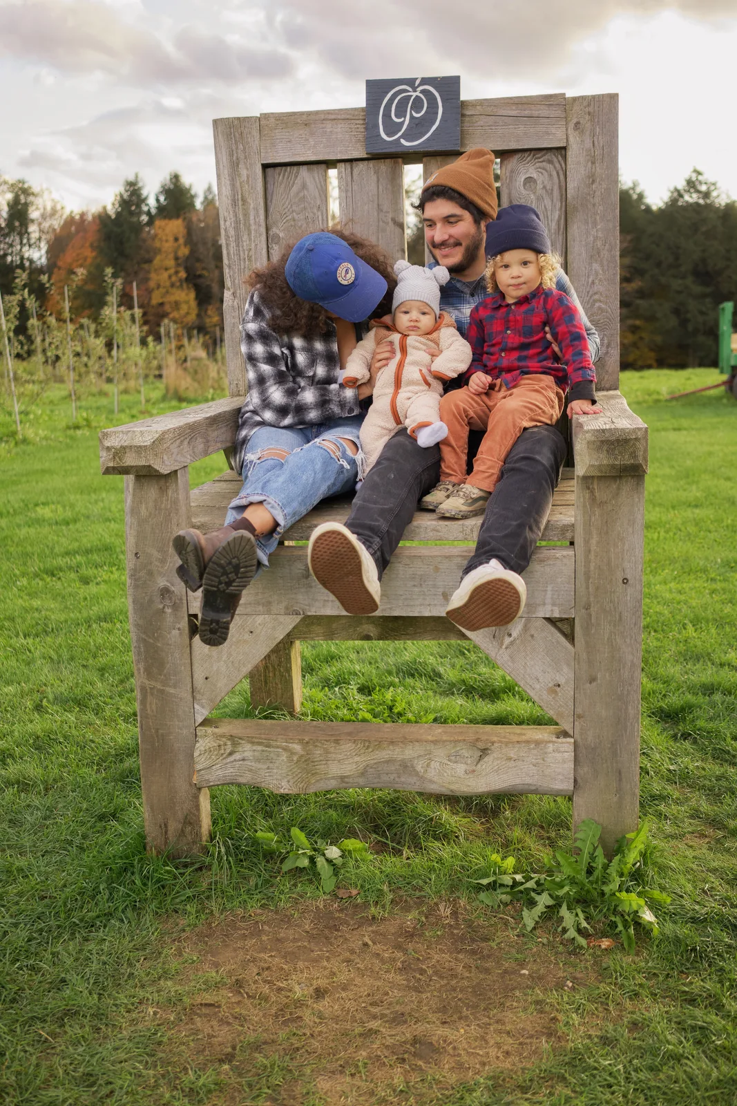 Four people seated on a large wooden chair in a grassy field with autumn trees