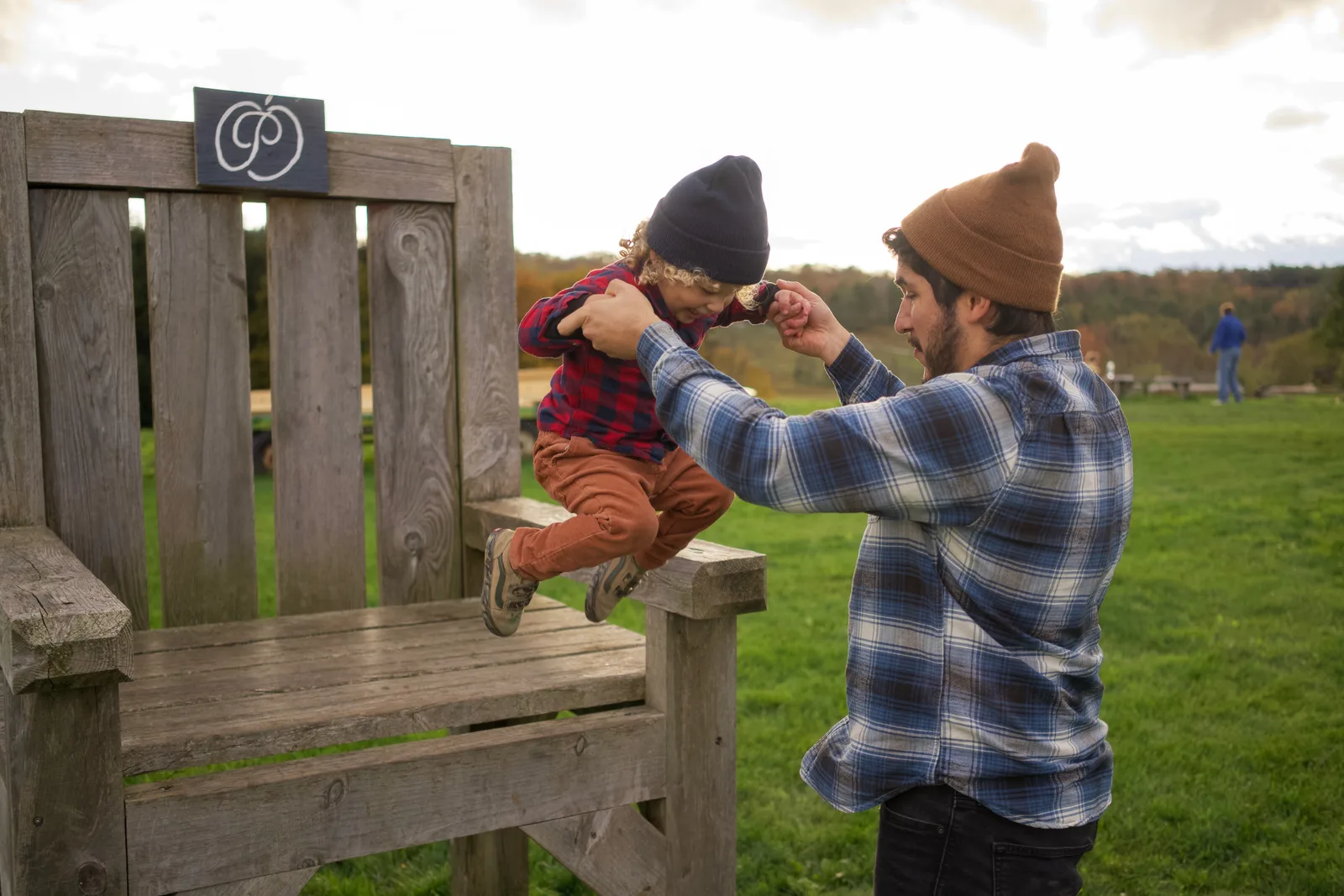 Adult in a brown beanie lifting a child in a black beanie onto a wooden chair